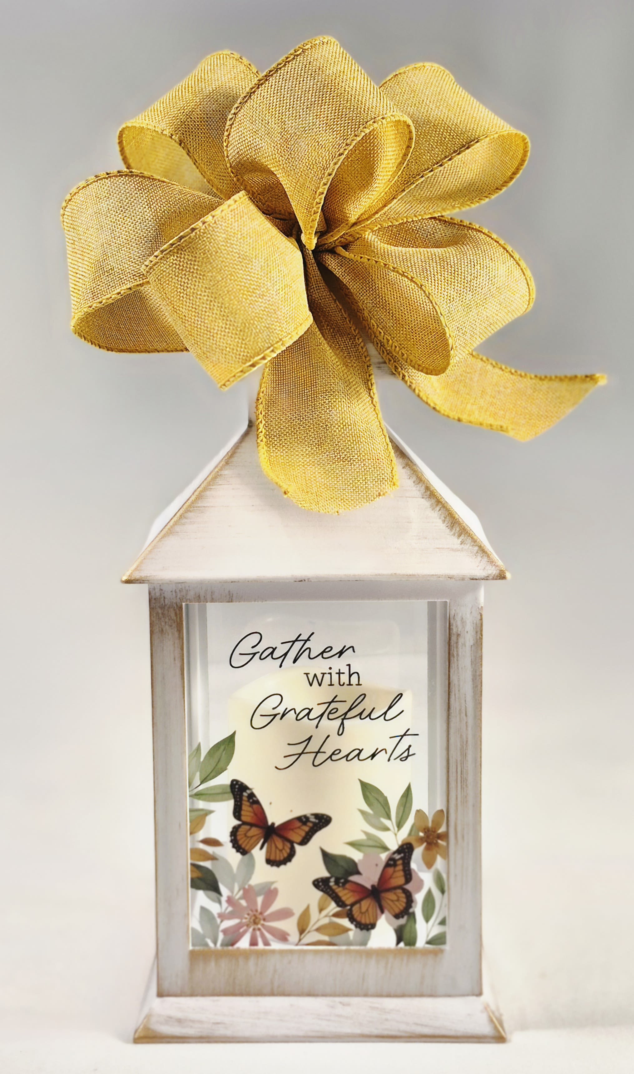 Gather with Grateful Hearts Lighted Lantern - A white decorative lantern topped with a yellow fabric bow sets a warm, inviting tone. This Gather with Grateful Hearts lighted lantern is designed beautifully with the printed sign on the front reading "Gather with Grateful Hearts" above a delicate butterfly and soft floral print to express gratitude for the person you're celebrating.  The lantern's rustic white finish paired with a warm metallic gold bow and mixed-color floral artwork blends farmhouse charm with polished detail. Black accents and the printed sign give clear seasonal messaging while keeping the piece versatile enough for mantel, entry table, or dining centerpiece. The included fabric bow and decorative hard goods make this a ready-to-display gift that sends a thoughtful, thankful message.  Choose this lantern when you want to make someone feel seen and appreciated at a holiday table or during a fall visit-it's the kind of home accent that quietly upgrades a room and prompts compliments. Dimensions and care are provided at checkout. H: 9" DIA: 4 "