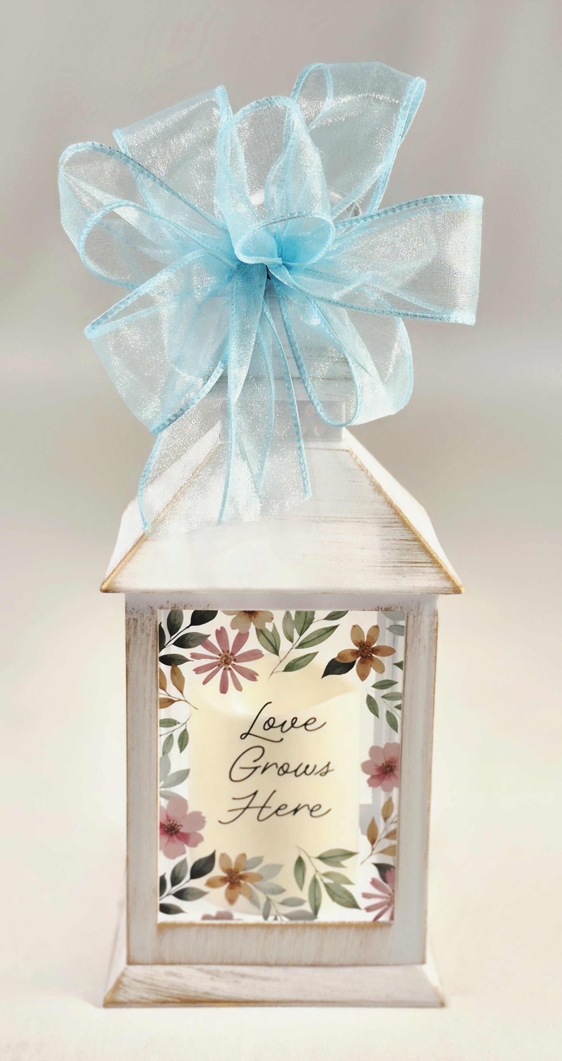 Love Grows Here Lighted Lantern - A distressed white decorative lantern topped with a sheer green bow creates a soft, welcoming focal point-perfect as a housewarming gift, a thank-you present, or an everyday floral accent for someone you love. This flower arrangement in a lighted lantern sends a calm, affectionate message: the printed front panel framed with floral art reads "Love Grows Here," so the recipient will feel thought of and warmly welcomed whenever they see it.  Inside the lantern, a gentle palette of white, blue, pink, and green florals and greenery complement the pastel finish for a fresh, cottage-style look. Overall dimensions are approximately 9" high with a 4" diameter, making it a compact yet charming piece for tabletops, entry consoles, or kitchen windowsills.  Ideal for celebrating a new home, offering thanks, or brightening someone's day, this arrangement doubles as keepsake home décor that can be displayed long after the flowers fade. The LED light inside provides a soft glow to showcase the florals and printed sentiment.