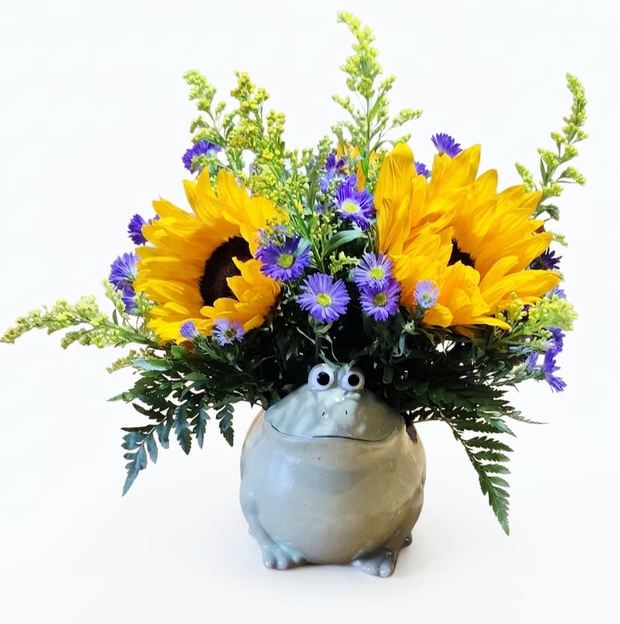 Hippity-Hop Sunflower Frog - Sunny sunflowers jump out of a whimsical ceramic frog vase, making this flower arrangement a playful pick-me-up for everyday celebrations or to brighten someone's day. Perfect as a birthday bouquet for a friend, a cheerful get-well surprise, or a just-because gift, the Hippity-Hop Sunflower Frog reads as lighthearted and fun while still feeling thoughtfully arranged.  Bright yellow sunflowers are paired with small purple aster-like blooms, airy yellow filler flowers, and touches of white and green that add freshness and contrast. The flowers sit in a rounded ceramic frog container with sculpted googly eyes, creating an immediate smile and a casual, upright silhouette that fits on a desk, windowsill, or kitchen counter.  Give this to the person you want to make feel seen and lifted-the effect is joyful and informal rather than formal or fussy. 
