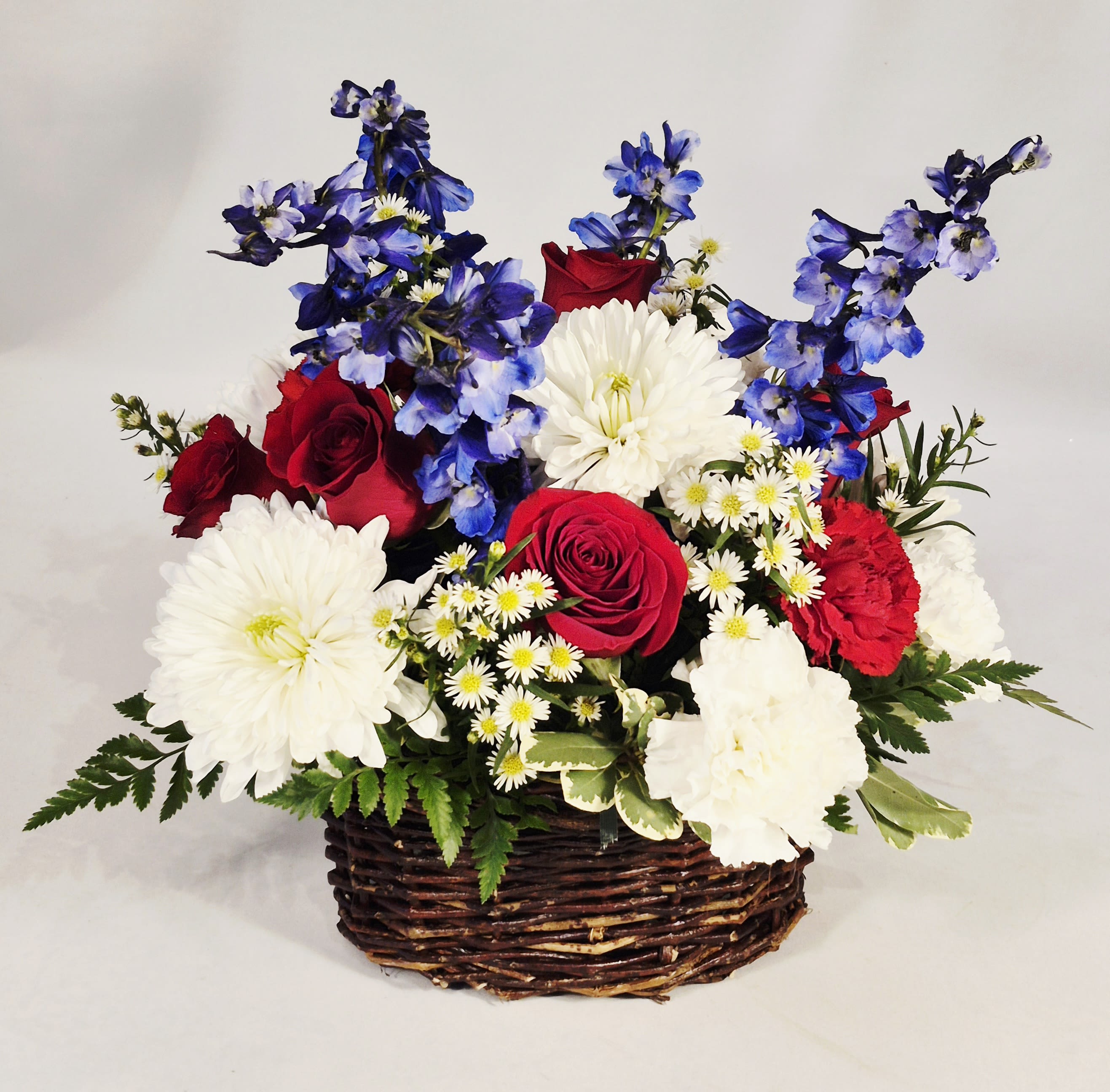 Red, White and Blue Basket  - Bright clusters of red roses set against large white blooms and vivid blue flowers make this Red, White and Blue basket an eye-catching choice for patriotic holidays, anniversaries, or to celebrate someone special. The arrangement reads as both festive and refined-the deep reds bring warmth, the crisp whites add elegance, and the pops of blue give it a cheerful, spirited finish that will make the recipient feel noticed and celebrated.  The mixed-color design is arranged low in a woven basket, measuring approximately 12 inches high and 10 inches wide, with roses, carnations, daisies and other seasonal blooms layered for texture and contrast. Small white daisy-like filler flowers soften the composition while the basket adds a rustic, homespun touch that complements both casual and more formal settings.  Perfect for gifts that say "thank you," "congratulations," "thinking of you", or is a wonderful sympathy arrangement ready to display on a table, mantle, or entryway where its colors can brighten the room. 