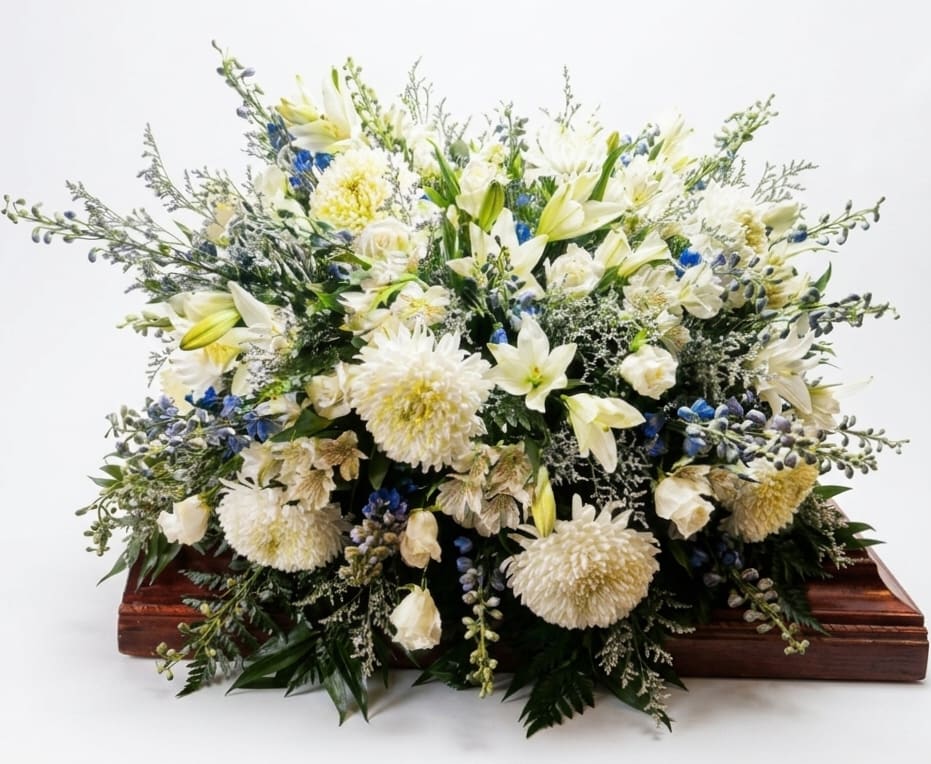 Bountiful Memories Casket Spray - White lilies, roses, chrysanthemums and alstroemeria form a serene, full spray with soft blue delphinium accents-an elegant casket spray for a funeral or memorial service. Placed on a wooden casket, the low, layered silhouette and trailing stems create a formal, composed presentation that reads as respectful and timeless for the person you're honoring.  This large arrangement is built on dense greenery-emerald palm and leatherleaf woven through clusters of limonium and other filler blooms-for depth and texture. Predominantly white blooms give a calm, luminous palette while the blue highlights provide a gentle, comforting contrast.  Designed to convey sympathy and support, this spray offers a meaningful way to let the person you're thinking of feel seen and remembered during the service. 