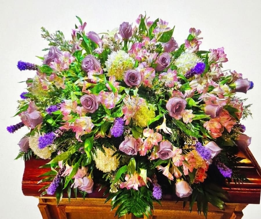 Lavender, Pink and White Casket Spray - Soft lavender, pink and white blooms come together in a graceful casket spray designed for sympathy and funeral services. This large, low arrangement makes an immediate visual statement across the casket with a traditional, formal silhouette that honors memories while offering gentle color and texture to mourners.  The spray is densely layered with lavender roses, pink alstroemeria, white chrysanthemums and purple accent flowers, finished with lush green foliage for depth. Its broad spread covers the top of the casket and slightly cascades over the edges, creating a full, continuous look from every angle. The combination of vivid pink, pale lavender and clean white provides a comforting palette that reads both soft and dignified.  Chosen for a loved one who deserves a tender, elegant tribute, this arrangement is especially fitting for memorial services where a warm, uplifting presence is desired. 