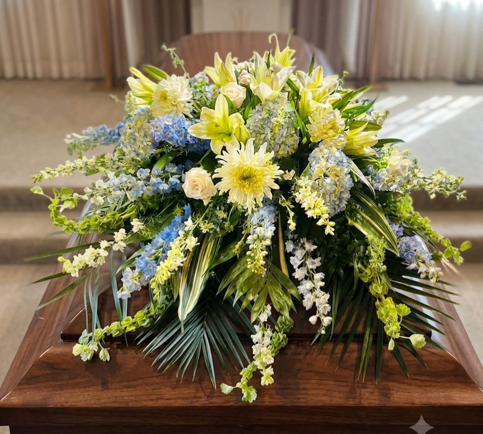 Seaside Memories Casket Spray  - Soft blue delphinium spikes and tinted blue hydrangea set a calm, coastal tone in this casket spray for sympathy and funeral services. Designed for someone who loved the water, the arrangement's white lilies and white roses form a dense, uplifting center while white daisy‑like blooms and bells of Ireland weave light texture throughout, creating a serene, memorial display.  This low, spray is built to drape gracefully across a casket: cascading blue and white delphinium-like spikes and trailing greenery lend layered movement and visual depth, with emerald palm and leatherleaf framing the composition. Conveys a comforting, thoughtful presence-an elegant way to honor a life with peaceful, seaside-inspired colors.