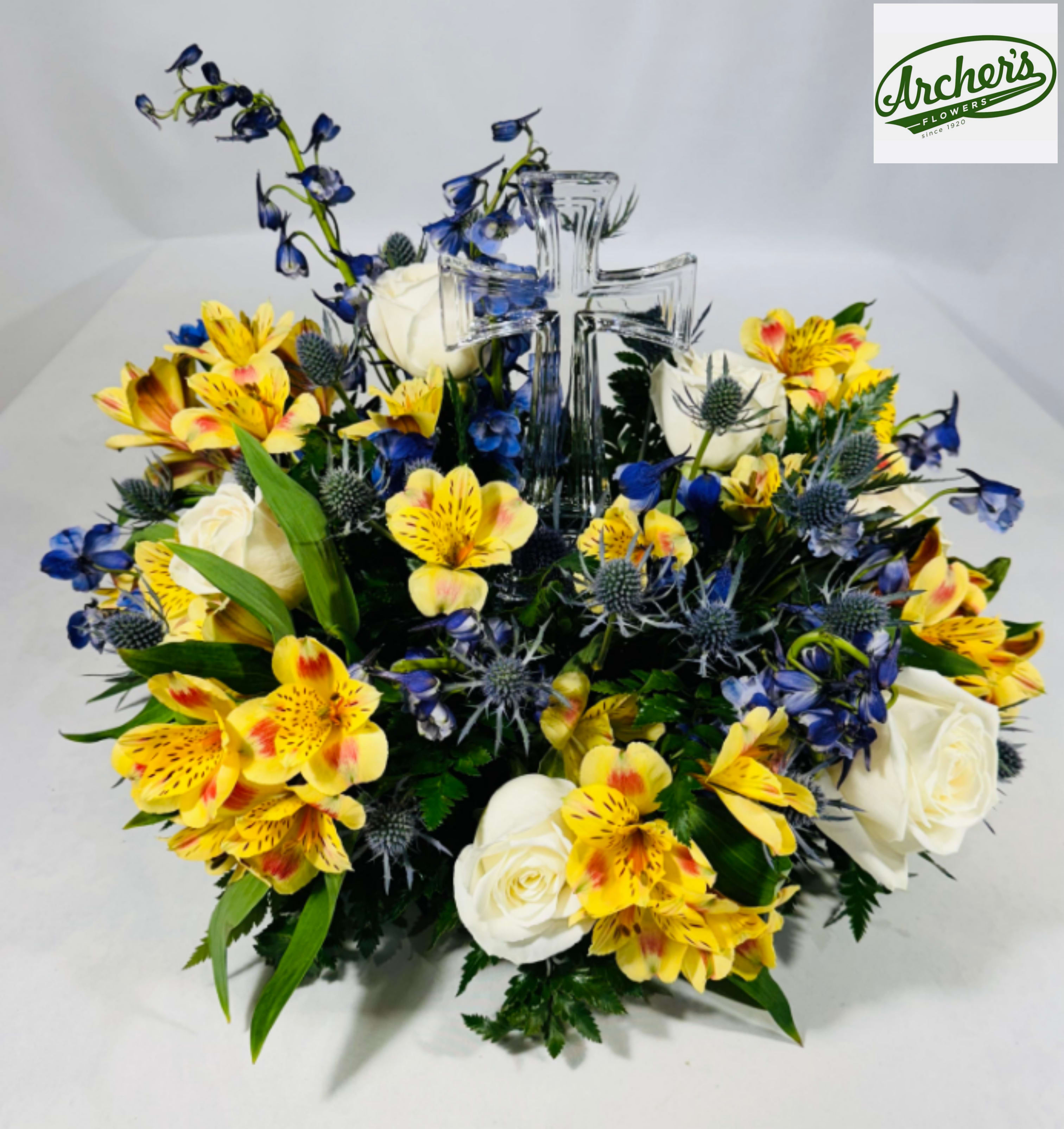Crystal Cross Fresh Arrangement - A clear crystal glass cross anchors this devotional sympathy arrangement, framed by a dense, low-and-wide spray of white roses, sunny yellow alstroemeria, vivid blue accents and mixed seasonal blooms-perfect as a sympathy or funeral tribute. The design reads both uplifting and reverent, making it an appropriate get-well gift for someone of faith or a thoughtful way to say you're thinking of them during a difficult time.  The composition is substantial on tabletops or memorial tables: layered white roses add softness and serenity, yellow alstroemeria brings warmth and hope, and the bright blue flowers provide crisp contrast against the transparent cross. Arranged compactly around the crystal glass cross, the flowers create a balanced, keepsake-ready presentation that feels both formal and comforting.  Ideal for sympathy, funeral services, Passover or Easter remembrances, or simply to let someone know you're thinking of them, this piece arrives ready to display. Dimensions: approximately 12" H x 14" W.