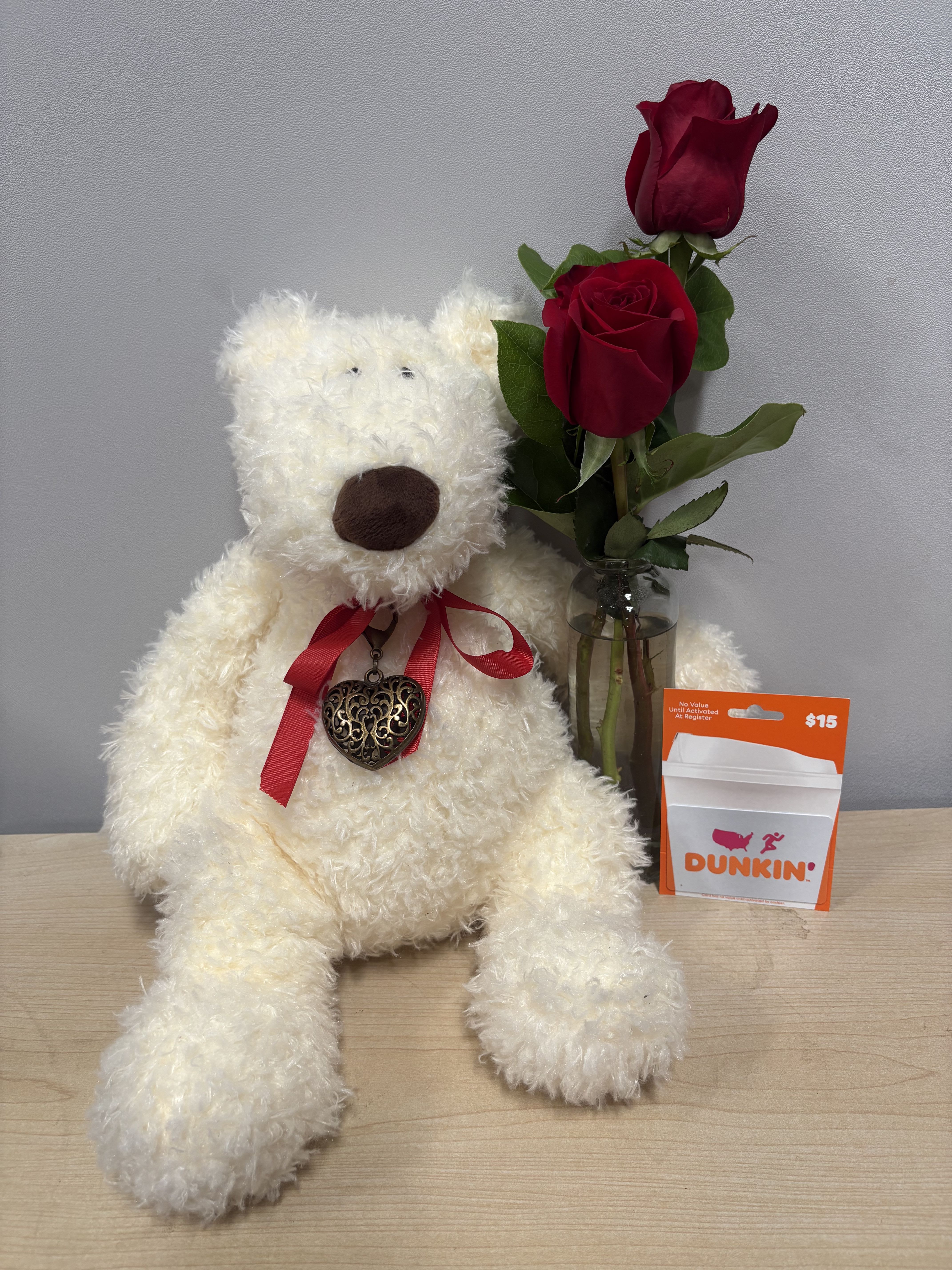 White teddy bear beside two red roses in a glass vase and a Dunkin' gift card