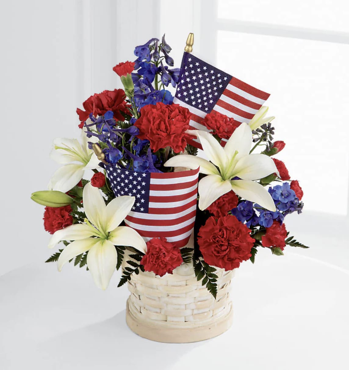 American Flag Basket - The American Glory Arrangement bursts with patriotic pride and heartfelt beauty. Blue delphinium, bright red carnations and brilliant white Asiatic lilies create a spectacular display arranged amongst American flags in a round small basket, creating a lovely way to celebrate the summer holidays.