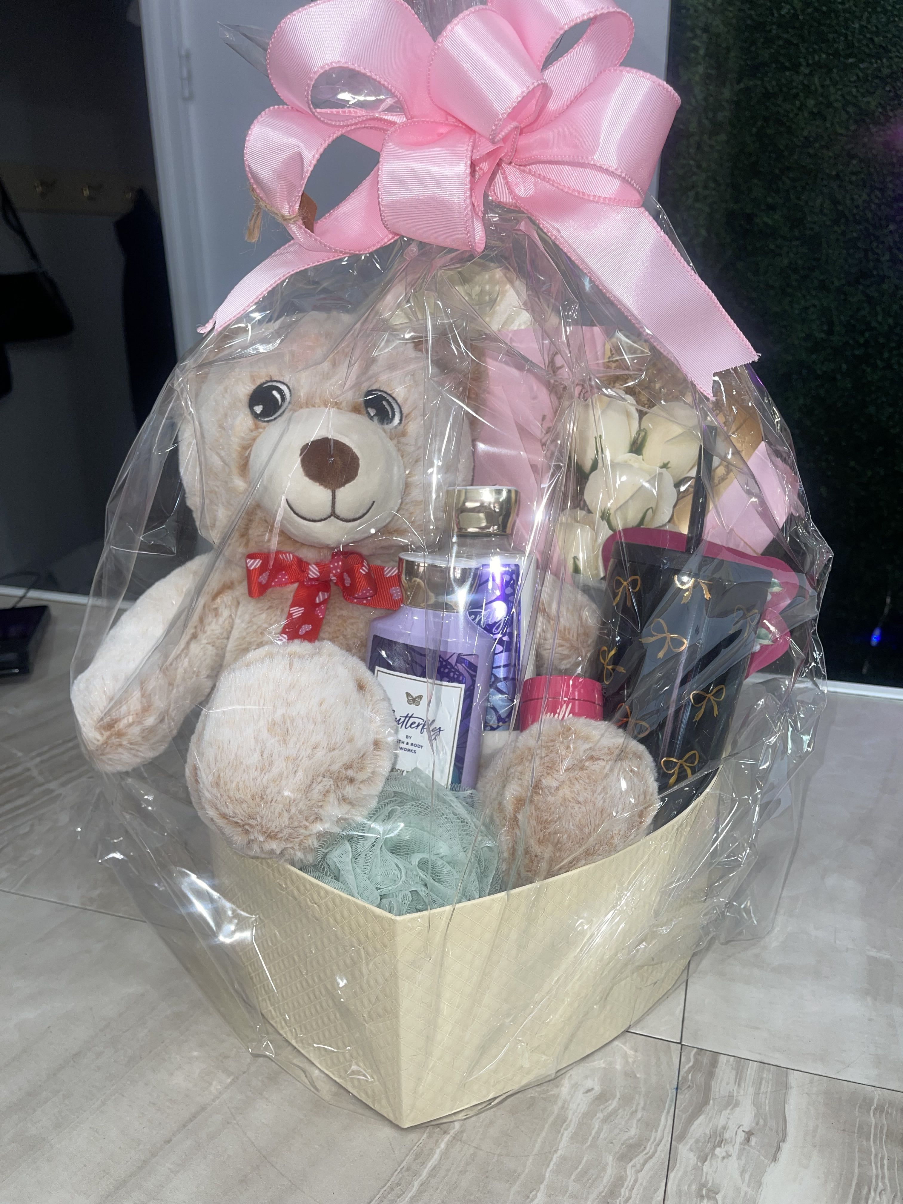 Gift Basket for Her -  Each basket arrives filled with assorted self-care and novelty items arranged in a woven or cream-colored container: bath and body products, hand cream, candle, perfume bottle, small cosmetic jars, a jewelry card with bracelet, a plush teddy or blue character plush, a white mug or decorative pouch, and other packaged treats-colors lean toward pink, white, lavender, blue and yellow for a gentle, feminine look. Wrapped with clear cellophane and ribbon, the presentation is cheerful and protective; choose the pink-accented basket for a classic feminine feel or the blue-handled option with a yellow ribbon for a playful twist.
