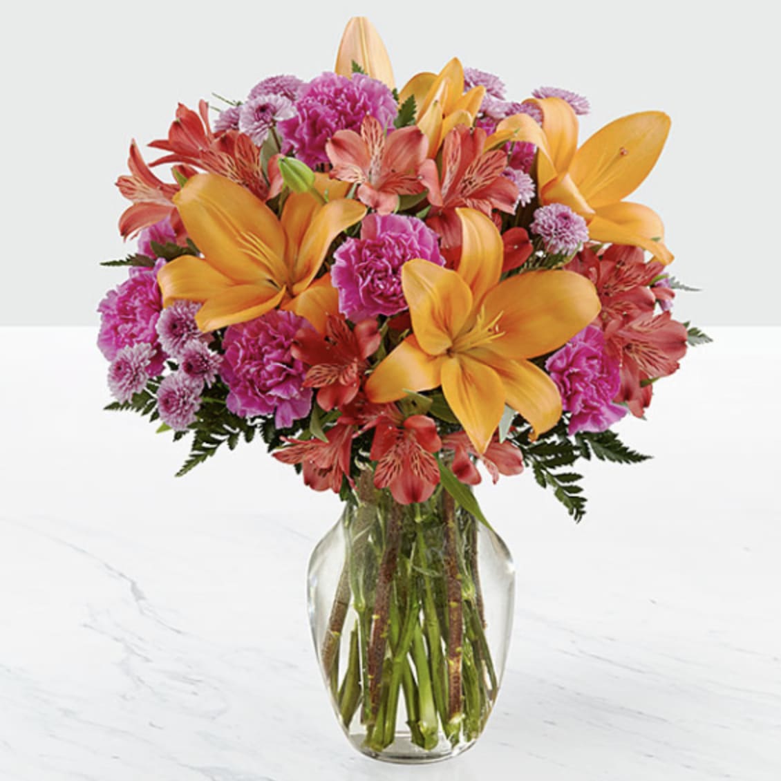 Orange Honey - Bright orange lilies stand proud above a lush, rounded bouquet in this cheerful flower arrangement-perfect as a birthday bouquet, congratulations gift, or just to brighten someone's day. The mix of orange, pink, and purple tones creates a lively, slightly tropical look that will make the person you're thinking of feel celebrated and seen.  This design pairs large orange lilies with coral and magenta alstroemeria, soft carnations, and textured chrysanthemums for a full, dense silhouette. The flowers are arranged tightly for lasting visual impact and come set in a simple clear glass vase that lets the vibrant blooms take center stage.  Made for everyday celebrations and happy occasions, this arrangement sends a message of warmth and joy without fuss. Vase care is straightforward-keep water fresh and trim stems as needed to extend longevity-and available in a classic size that fits a table, desk, or entryway display.