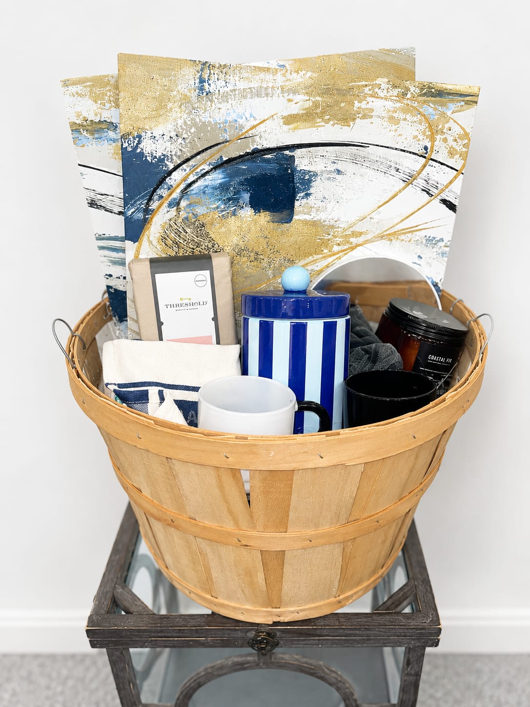 Homewarming Basket - A fresh, gift-ready housewarming basket that delights at first glance:  gift-basket  coastal-chic with a blue-and-white lidded ceramic jar, coordinating mug, folded towel and assorted candles and packaged treats, so the person you're celebrating will feel welcomed and cared for.  It is thoughtfully styled for everyday display and make a warm gift for housewarming, thank-you, or birthday moments. The wooden tub basket is compact and sculptural - easy to set on an entry way - thanks to the decorative panels at the back, creating an immediate, styled impression.  comes finished with ribbon, easy gifting; include any delivery notes at checkout so the recipient can enjoy it right away.