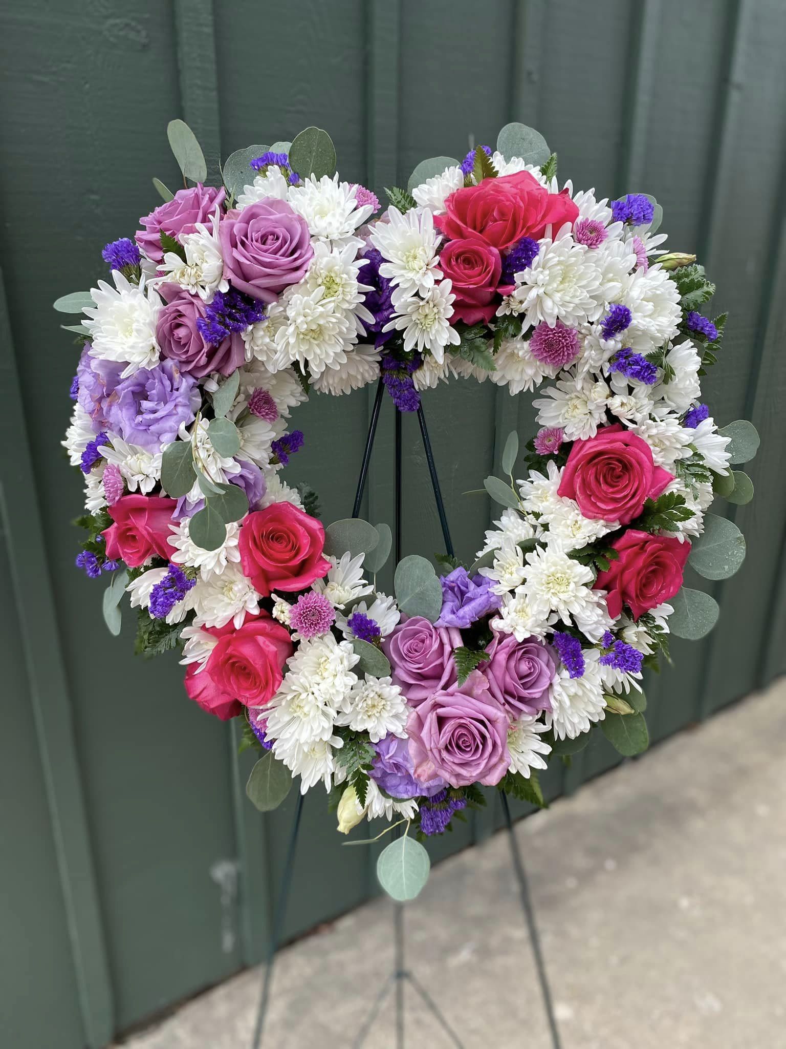 Heart-shaped wreath of pink and lavender roses with white mums and purple accents on a standing easel.