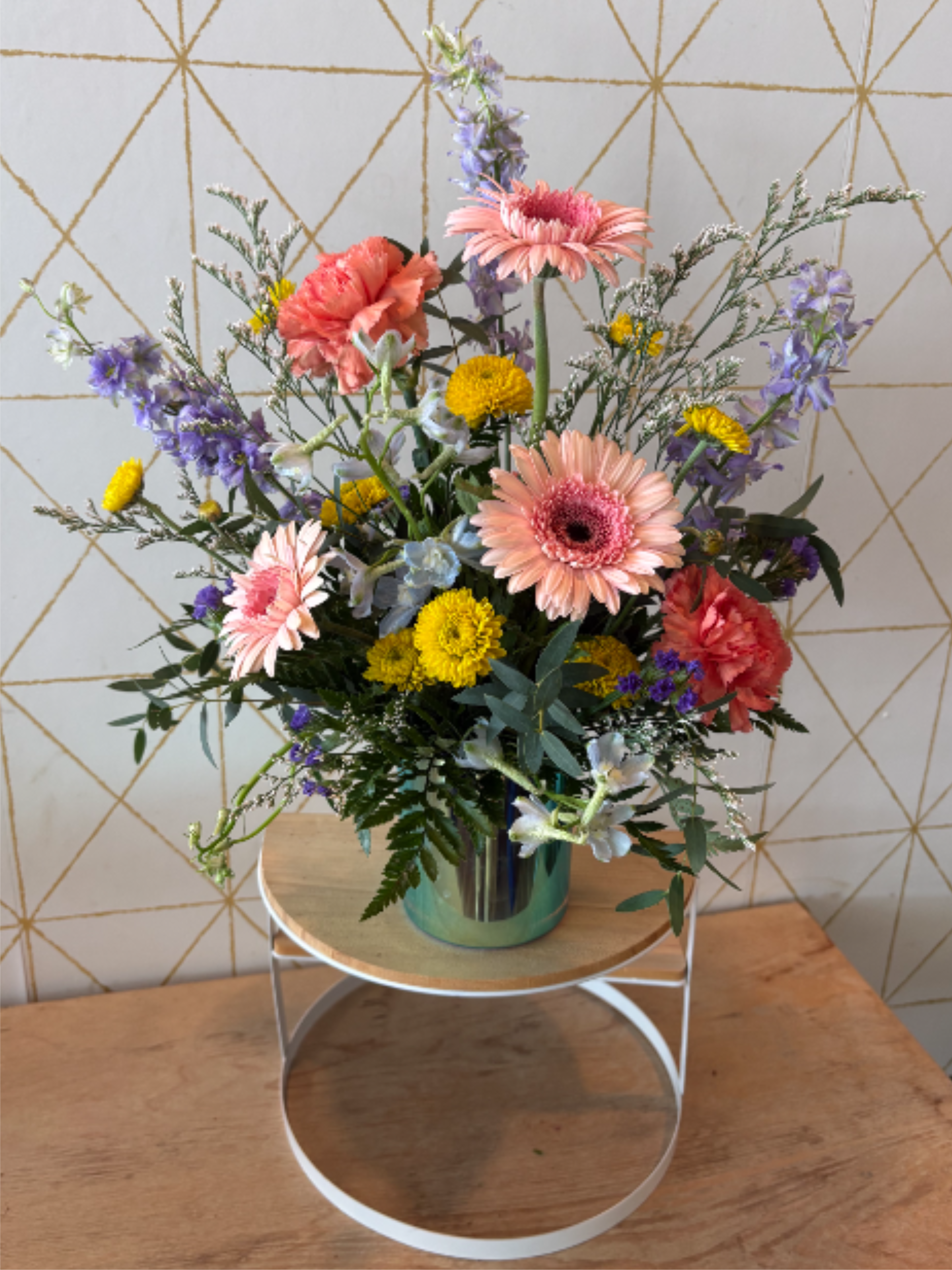 The Kourtney - Playful, polished and full of personality, The Kourtney features blush gerbera daisies, coral carnations, lavender larkspur, sunny button mums and textural filler and greenery accents arranged in an irridecent vase.  Perfect for celebrating life's sweetest moments or simply brightening someone's day.