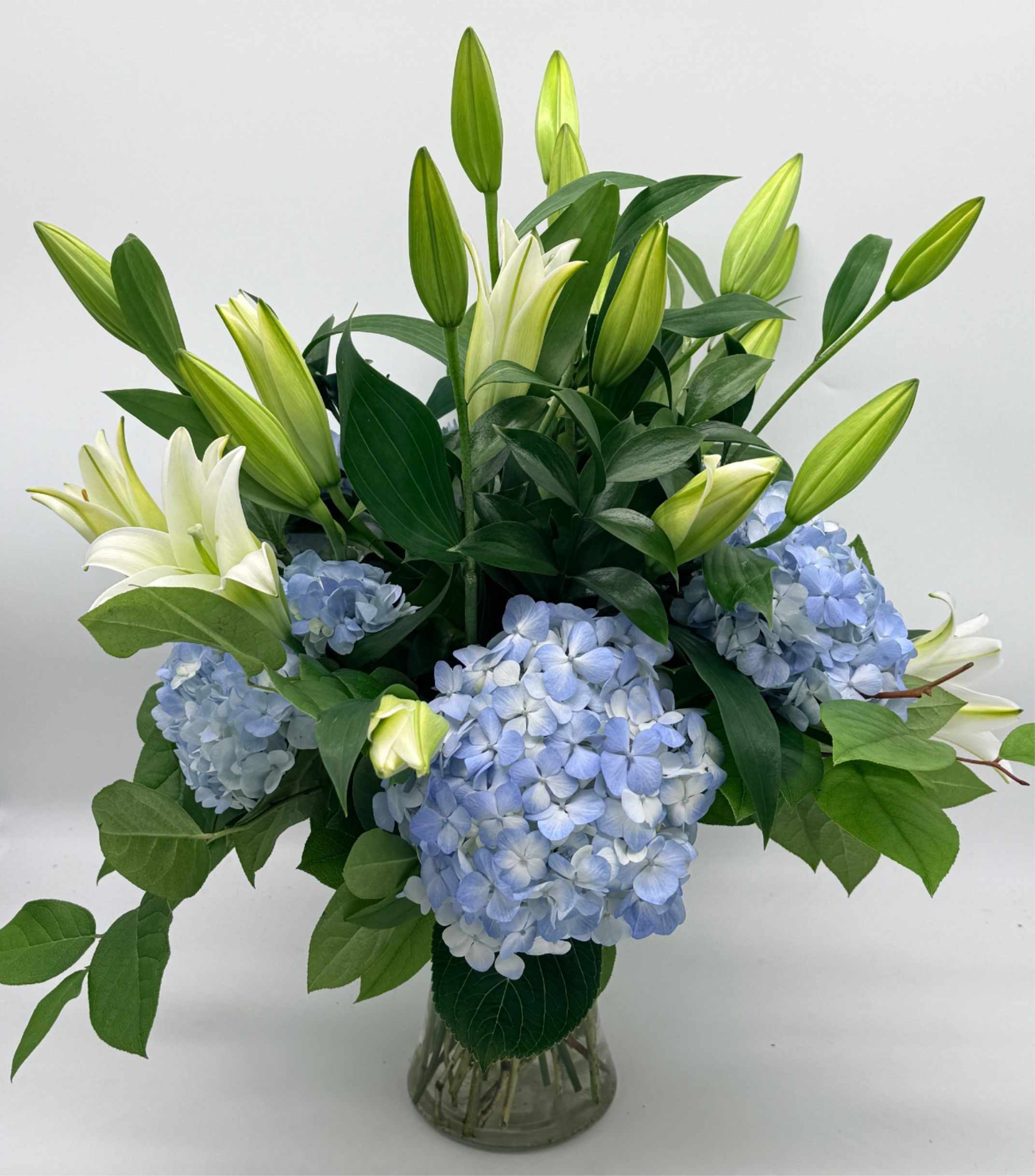 Coastal Elegance - Soft blue hydrangeas and pristine white lilies come together in this fresh, airy arrangement inspired by the calm of the coast.  Arranged in a clear glass vase with lush greenery, it’s perfect for birthdays, sympathy, thank you gifts, or everyday moments that call for something serene and sophisticated. 