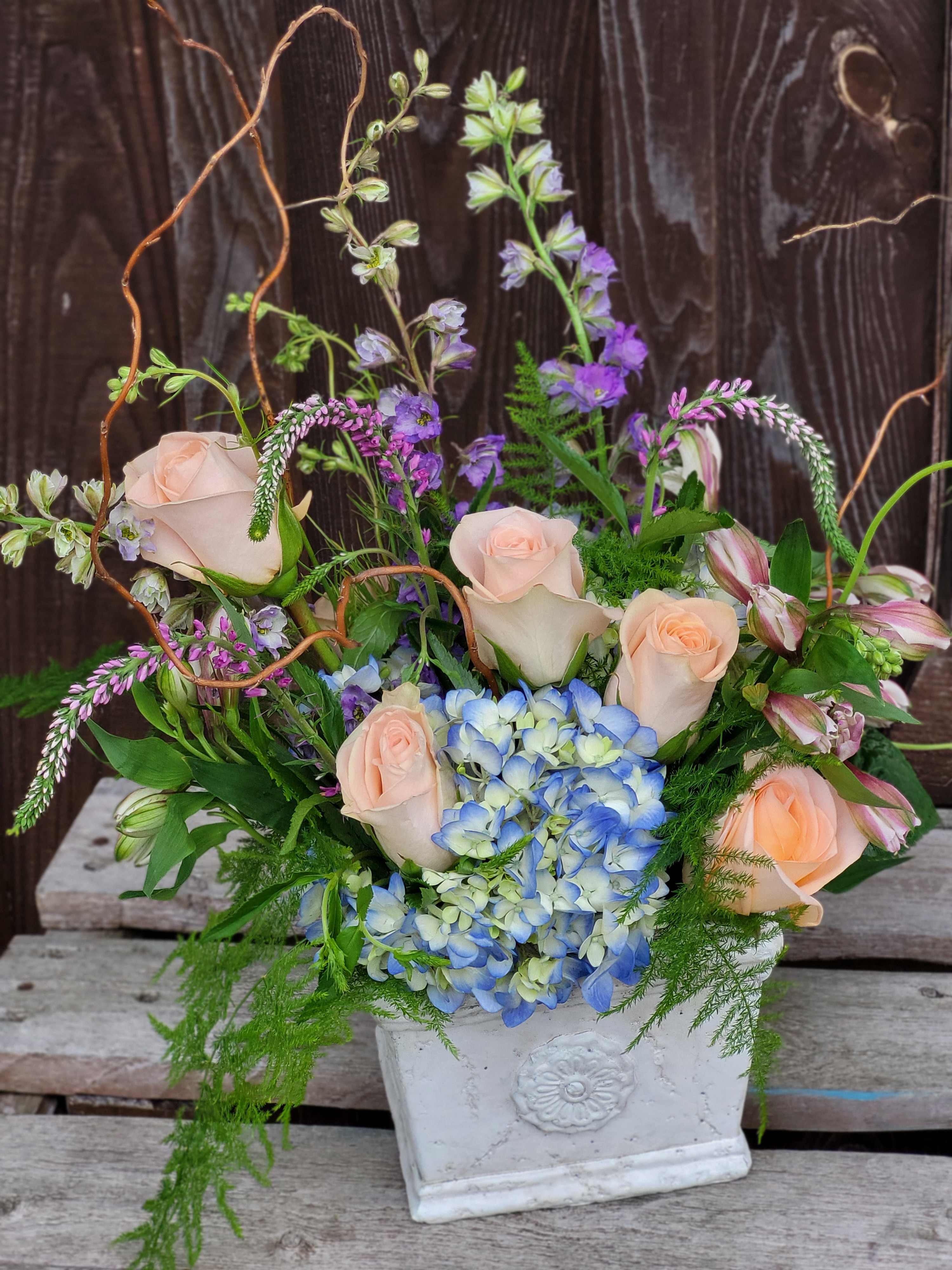 Mixed bouquet with pale roses, blue hydrangea, and purple blooms in a white vase