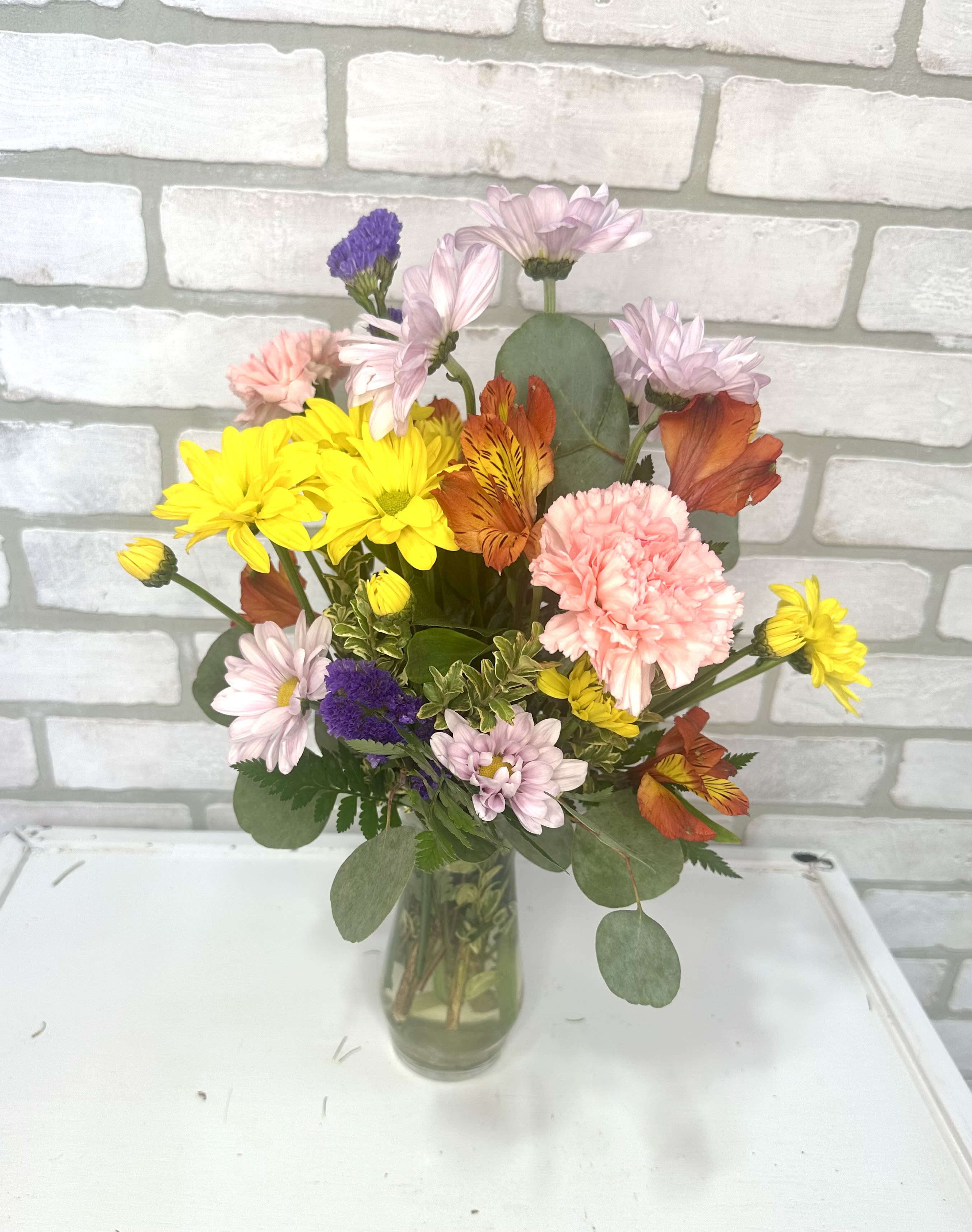 Mixed bouquet of yellow daisies, pink carnations, and orange blooms in a clear glass vase