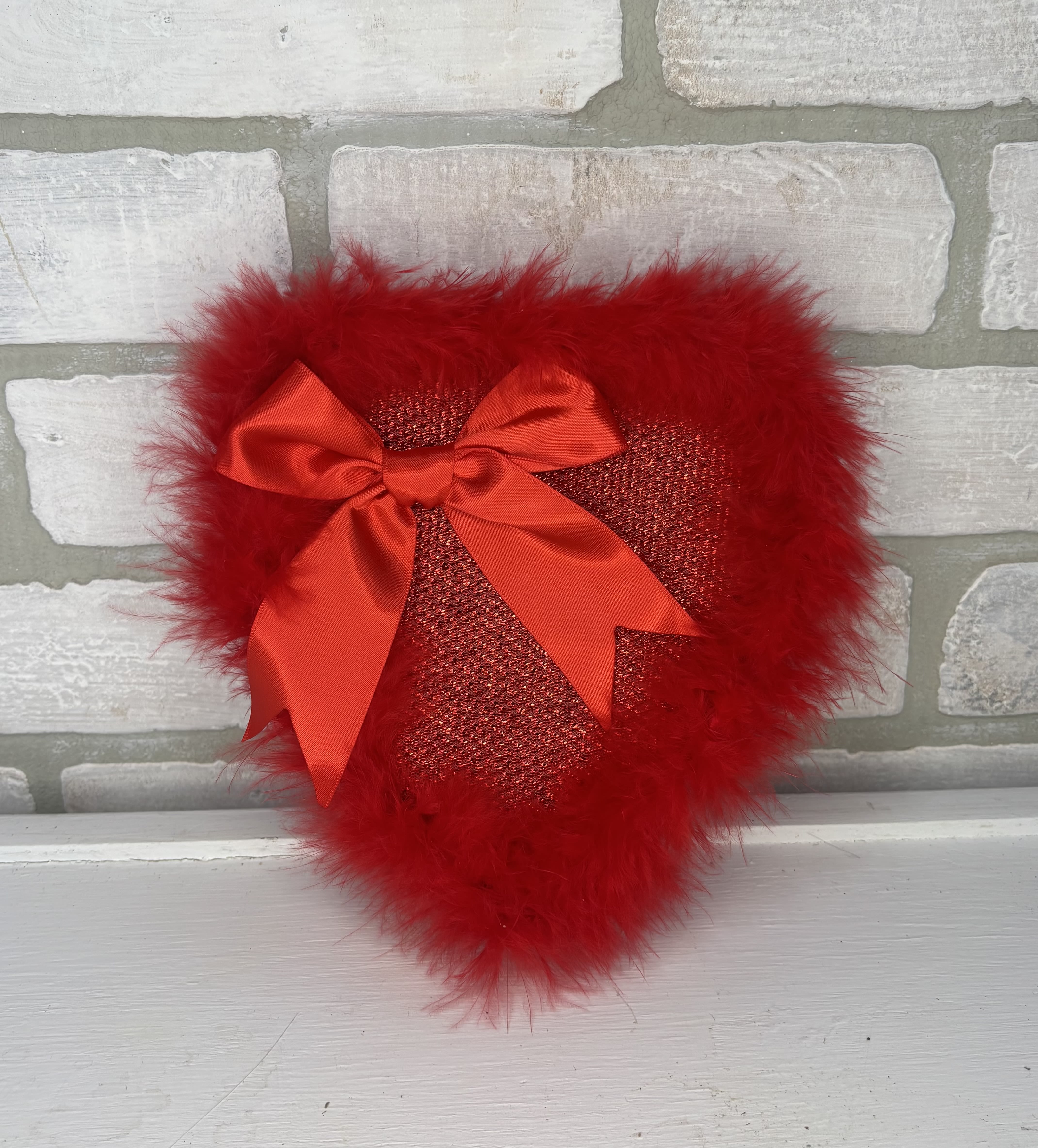 Red heart-shaped decorative pillow with feather trim and satin bow on a light brick background