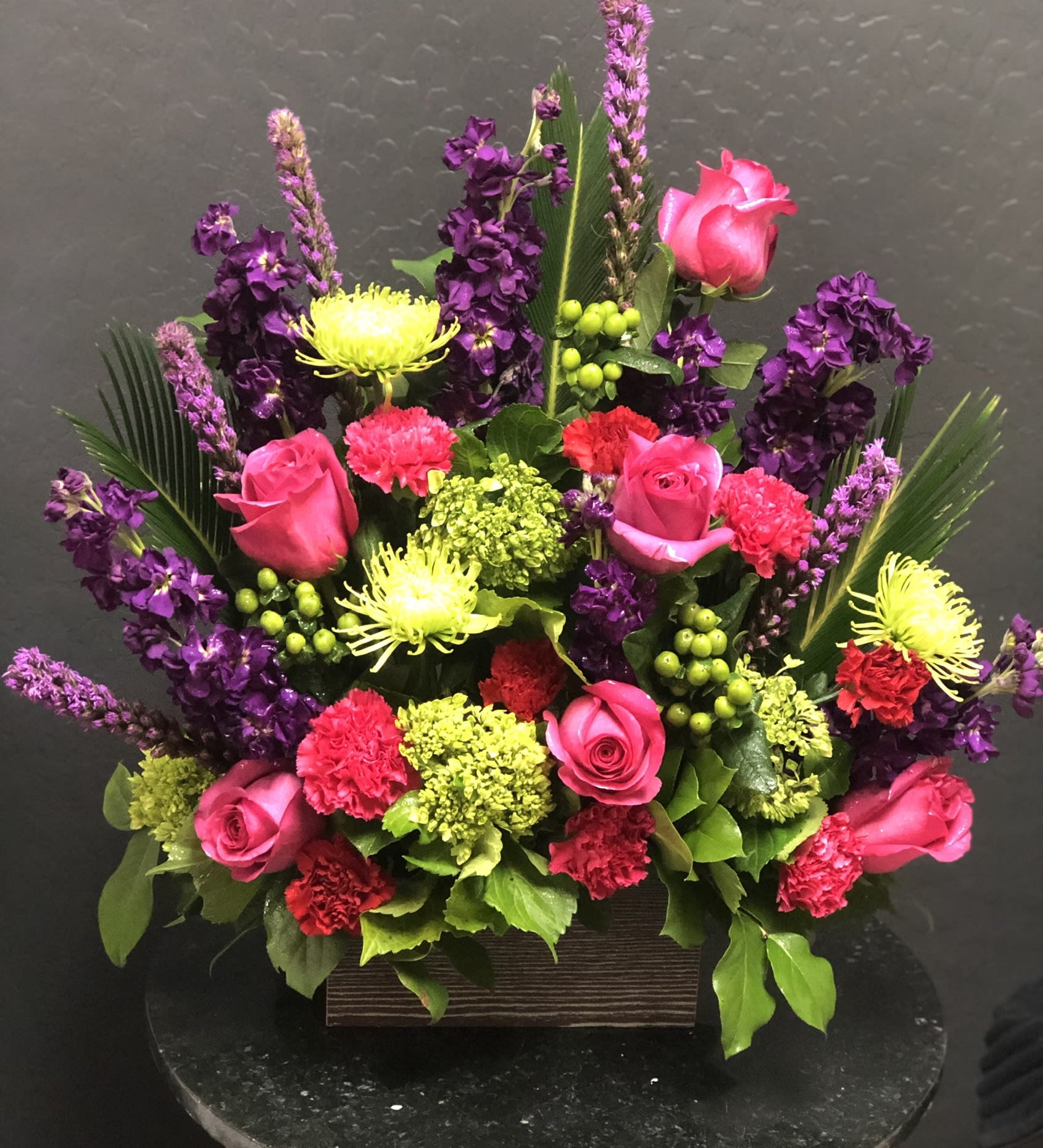 The FTD Warm Embrace Arrangement - A lovely way to say “farewell ” this bright beautiful arrangement expresses your sympathy with color texture and floral artistry while comforting grieving friends and family. This handcrafted bouquet is handcrafted by a local FTD florist and makes a vibrant statement with hot pink roses pink and fuchsia carnations purple stock green spider chrysanthemums mini-hydrangea and hypericum berries all accented with sword fern and salal in a low rectangle container of silvery metallic plastic.