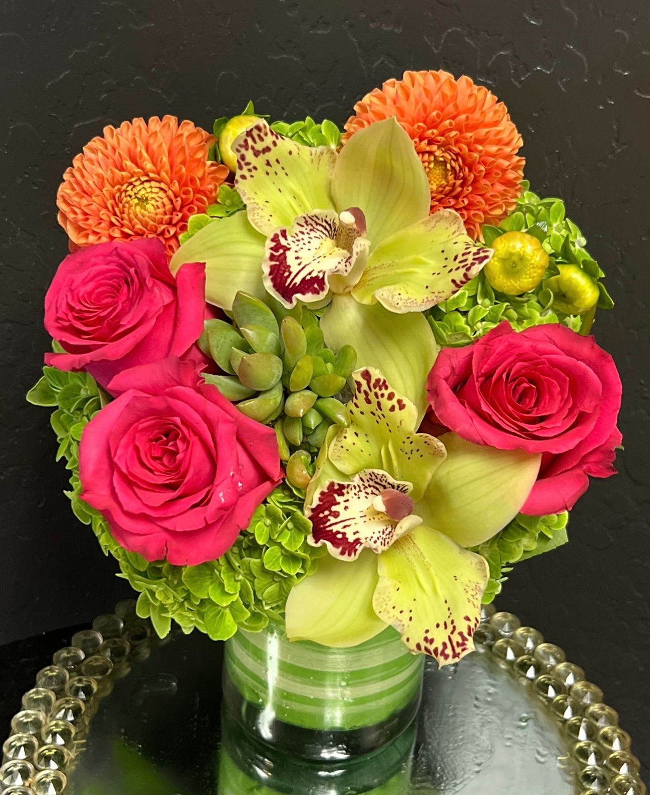 Pink roses and green orchids arranged with orange dahlias in a glass vase.