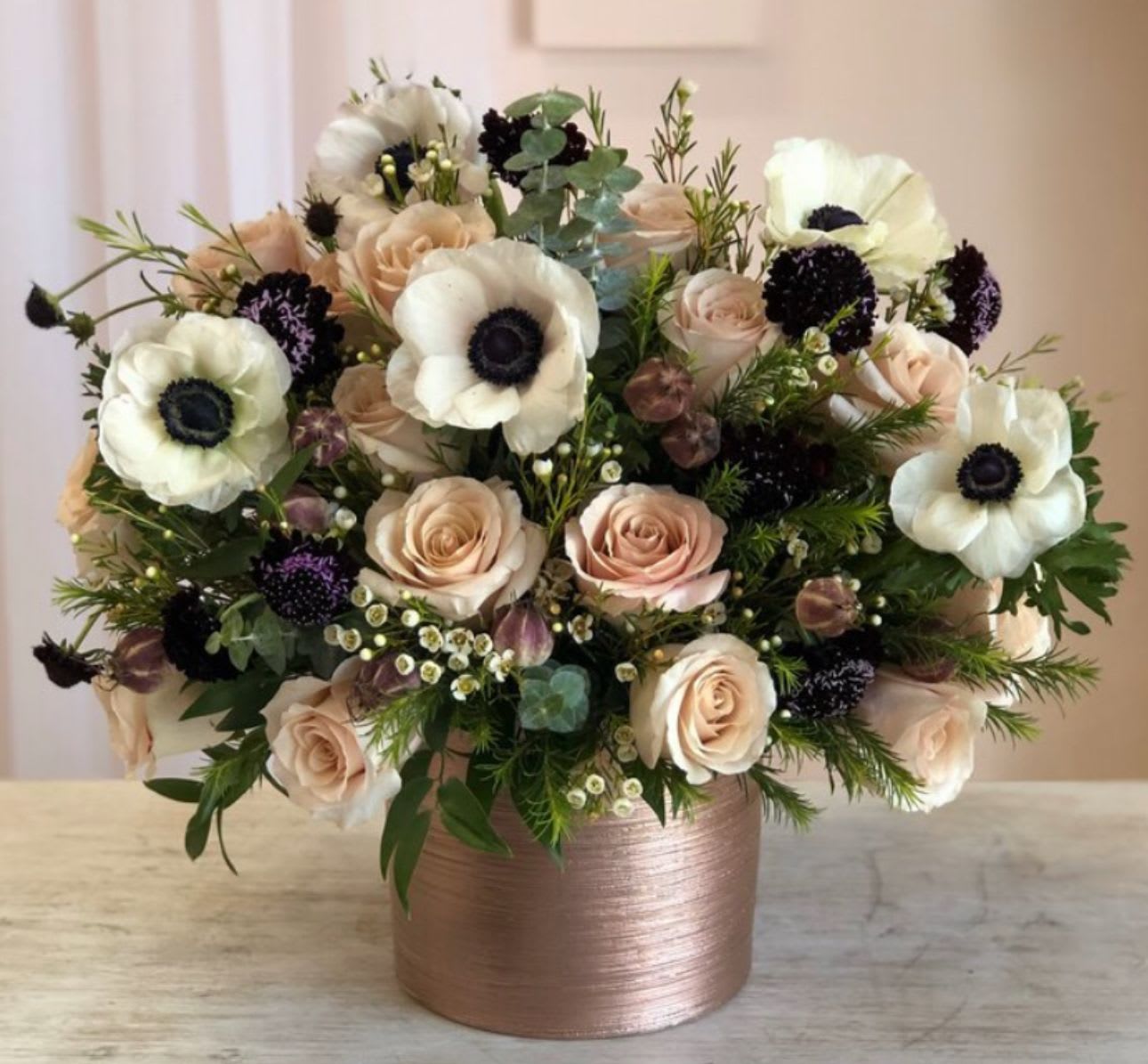 Arrangement of blush roses, white anemones, and dark blooms in a rose gold cylinder vase