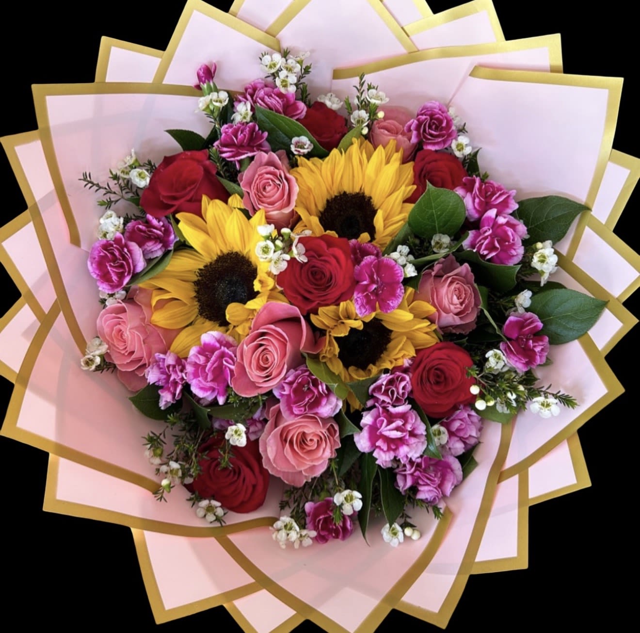 Sunflower and Rose Wrapped Bouquet  - Bright yellow sunflowers set a joyful tone against deep red and pink roses in this sunflower and rose wrapped bouquet-perfect as a birthday bouquet, anniversary gift, or a cheerful way to say "congratulations." The bold contrast of warm gold and rich crimson reads both romantic and celebratory, so the person you're thinking of will feel truly seen and delighted when they receive it.  The arrangement layers long-stemmed roses, sunflowers, and clusters of pink carnations with small white filler flowers and airy greenery for soft texture and fullness. Wrapped in decorative paper-options shown include a burgundy wrap with gold edging and a pale pink wrap with gold trim-and tied with a large matching ribbon, the presentation is polished and gift-ready. Stems are hand-tied for easy vase placement; simply trim the stems, change the water every couple of days, and keep away from direct heat to help blooms last longer.