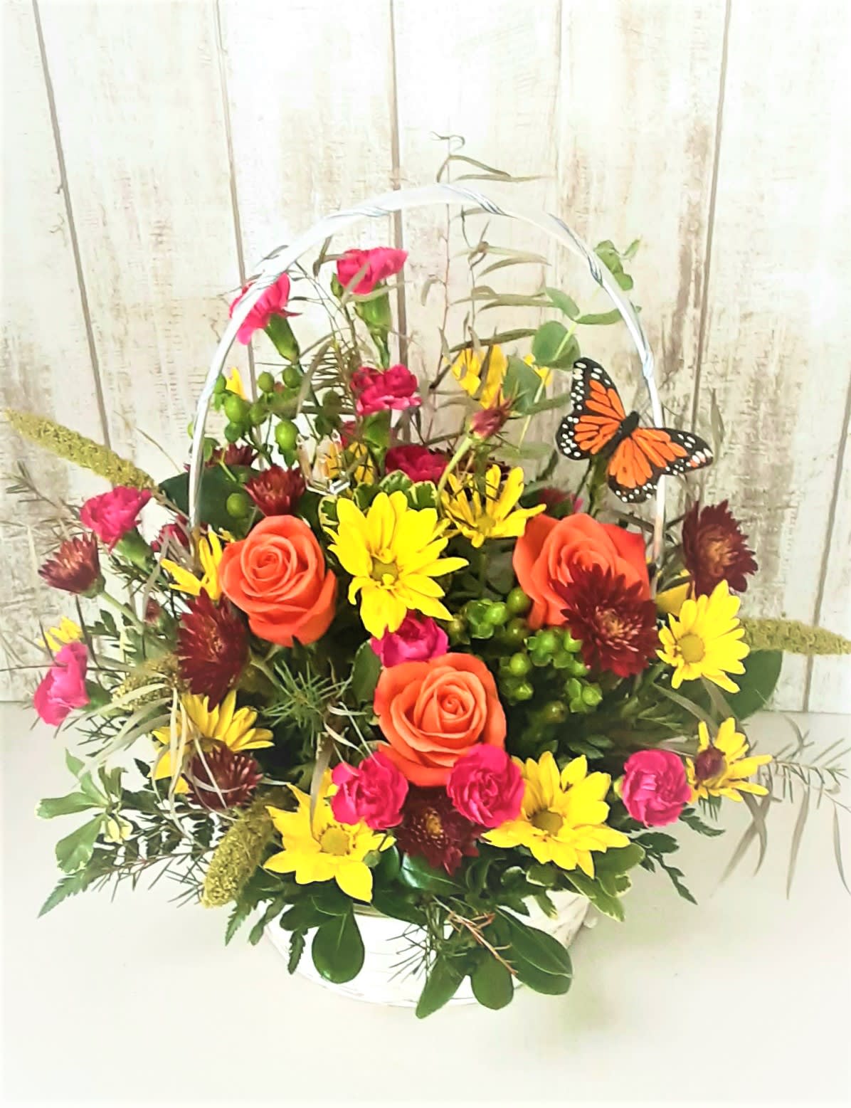 Basket arrangement of orange roses, yellow daisies, and pink flowers with a butterfly pick