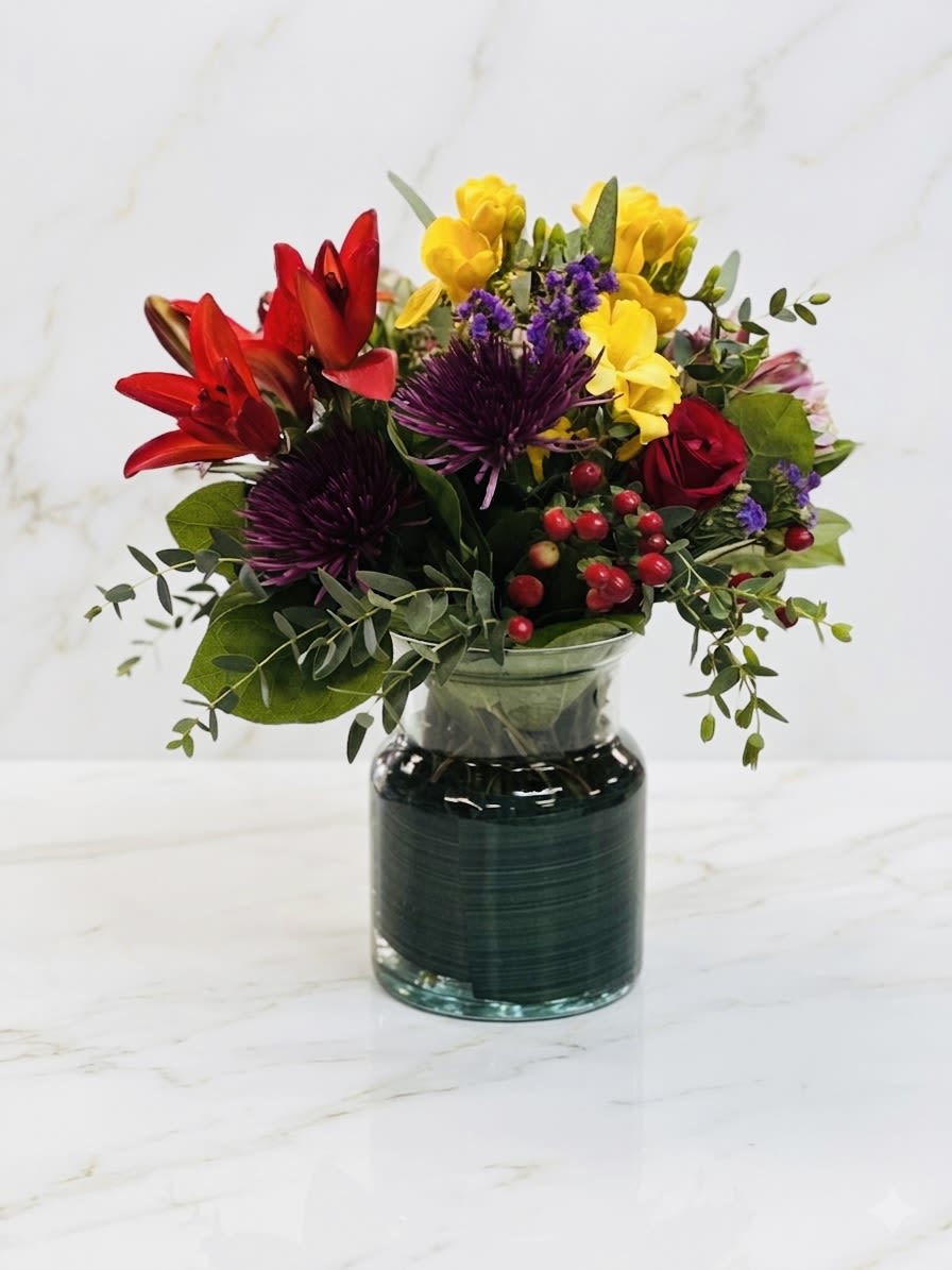 Bright mixed bouquet with red lilies, yellow blooms, purple mums and red roses in a clear glass vase