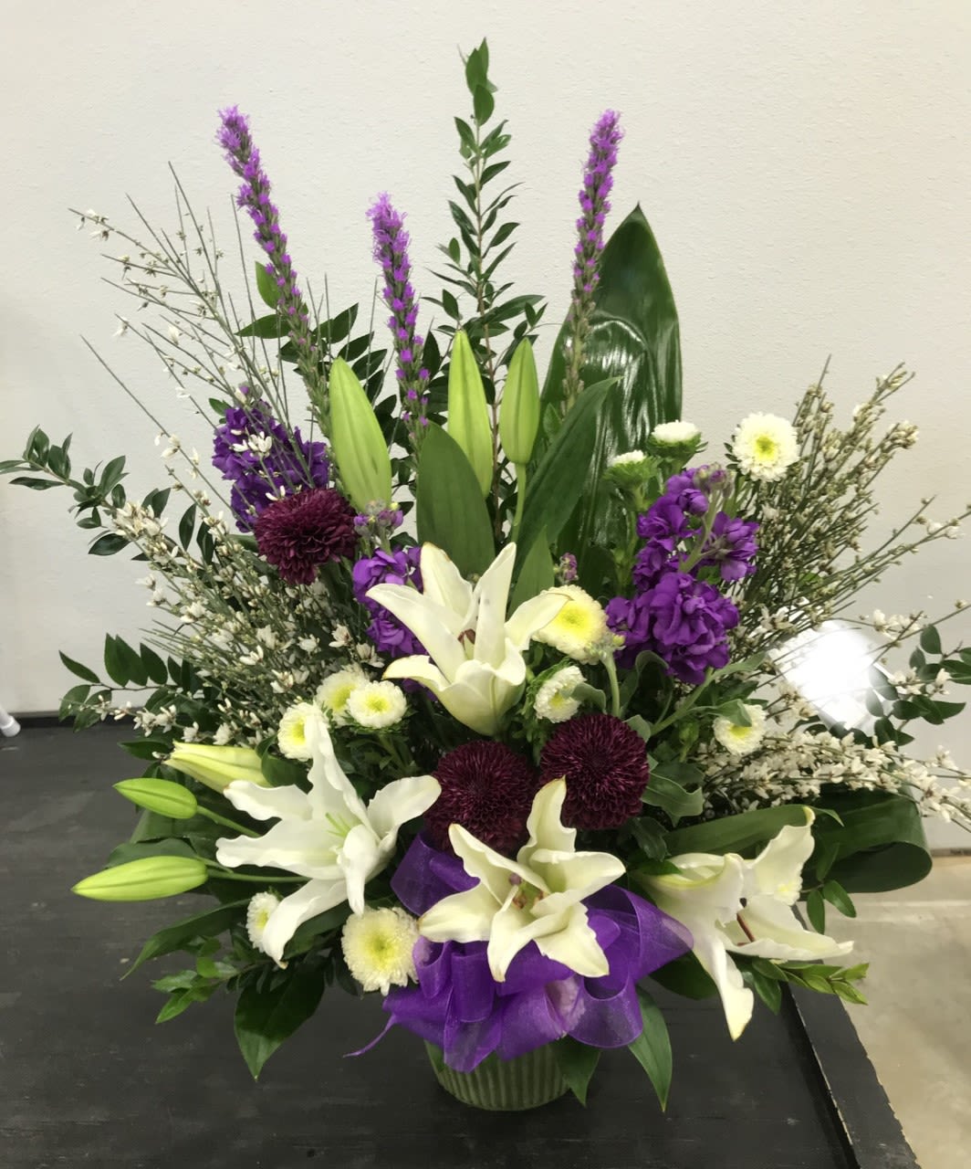 Tall floral arrangement with white lilies, purple blooms, and a purple ribbon in a vase