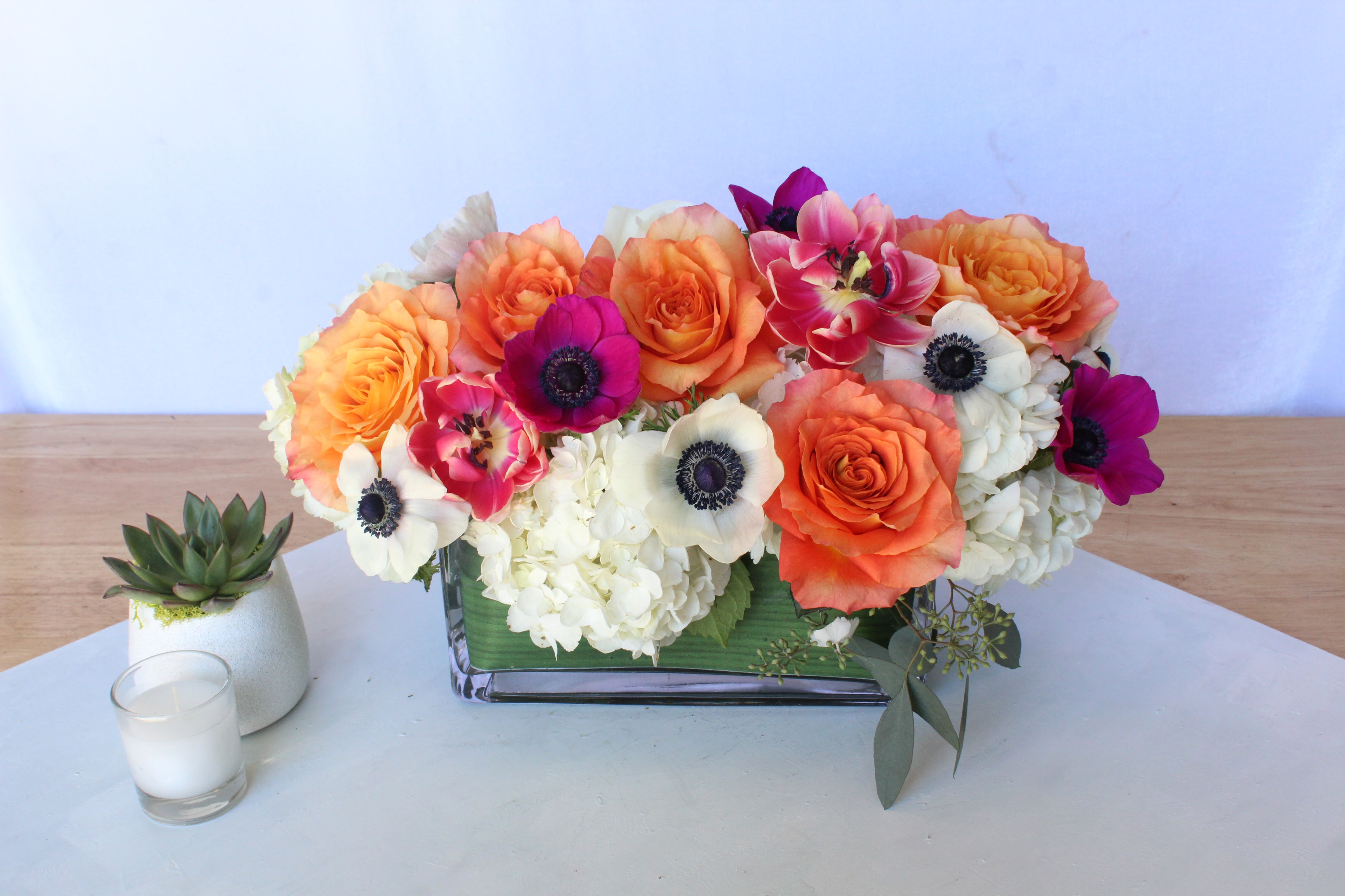Orange and white floral arrangement in a rectangular glass vase beside a small succulent and candle