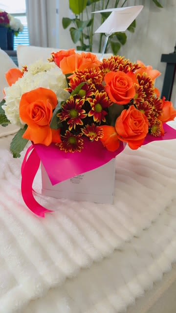Orange roses and burgundy daisies in a white gift box with a pink ribbon.