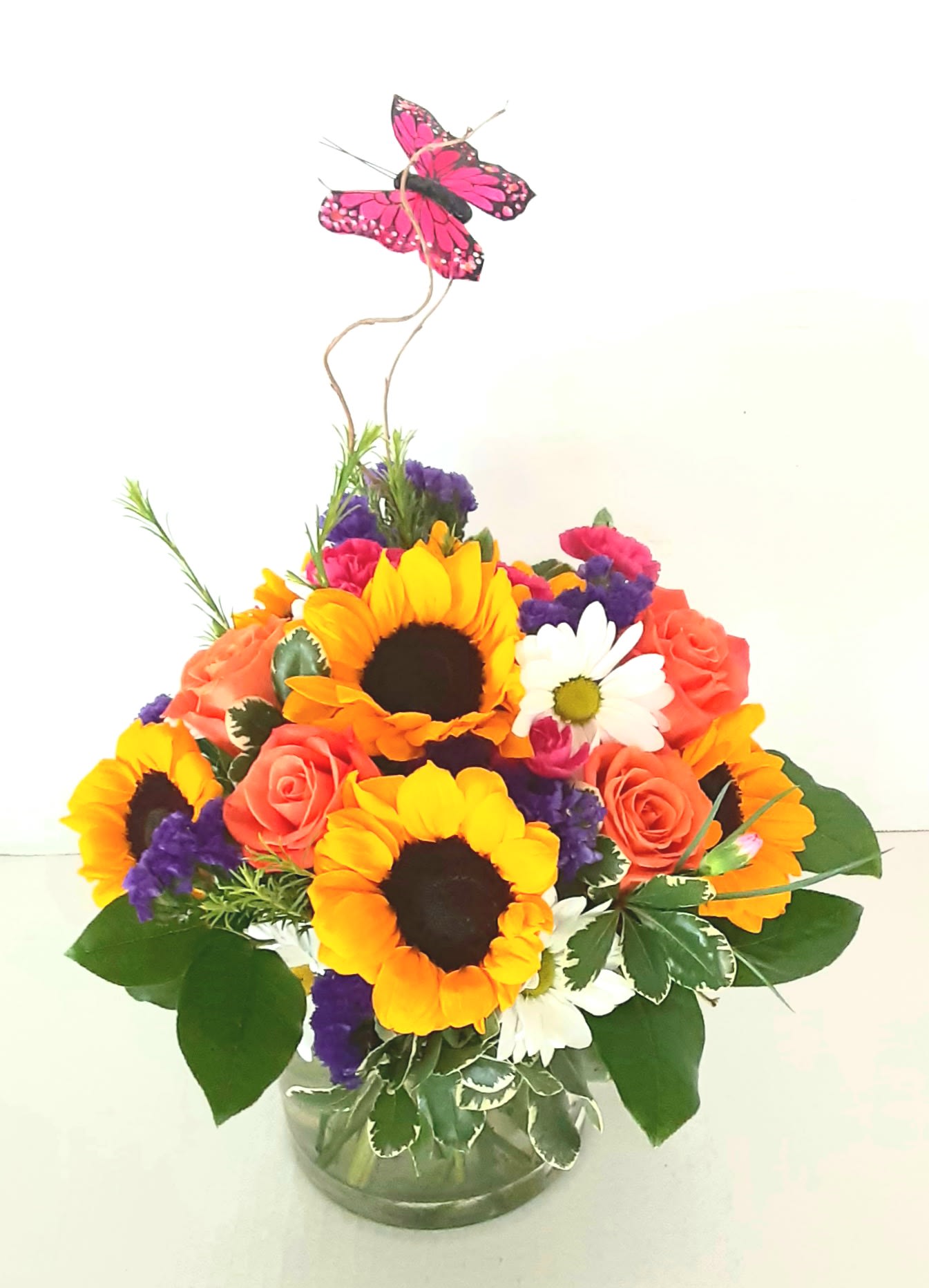 Mixed bouquet with sunflowers, roses, and daisies in a glass vase topped with a butterfly