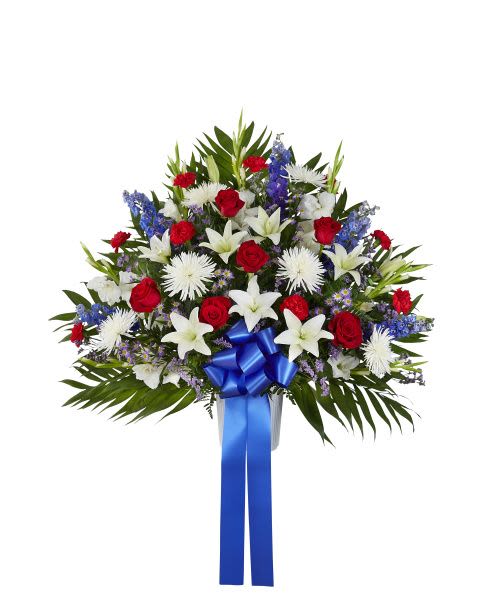 Fan-shaped red, white, and blue arrangement with roses, lilies, mums, and a blue ribbon in a white urn.