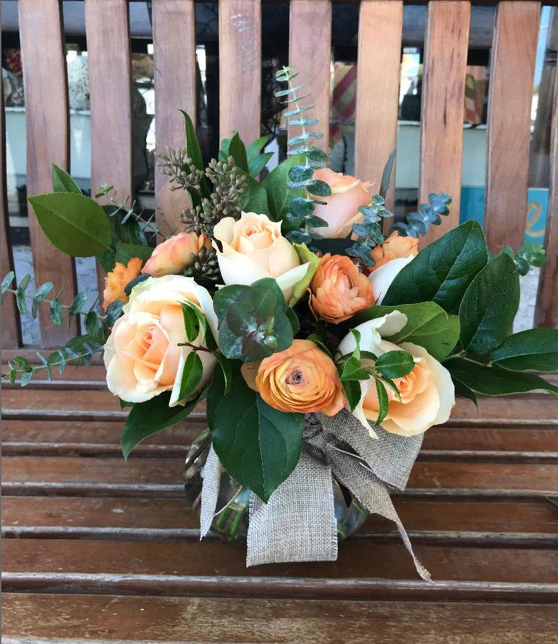 Peach roses and orange blooms arranged in a clear glass vase with a burlap bow