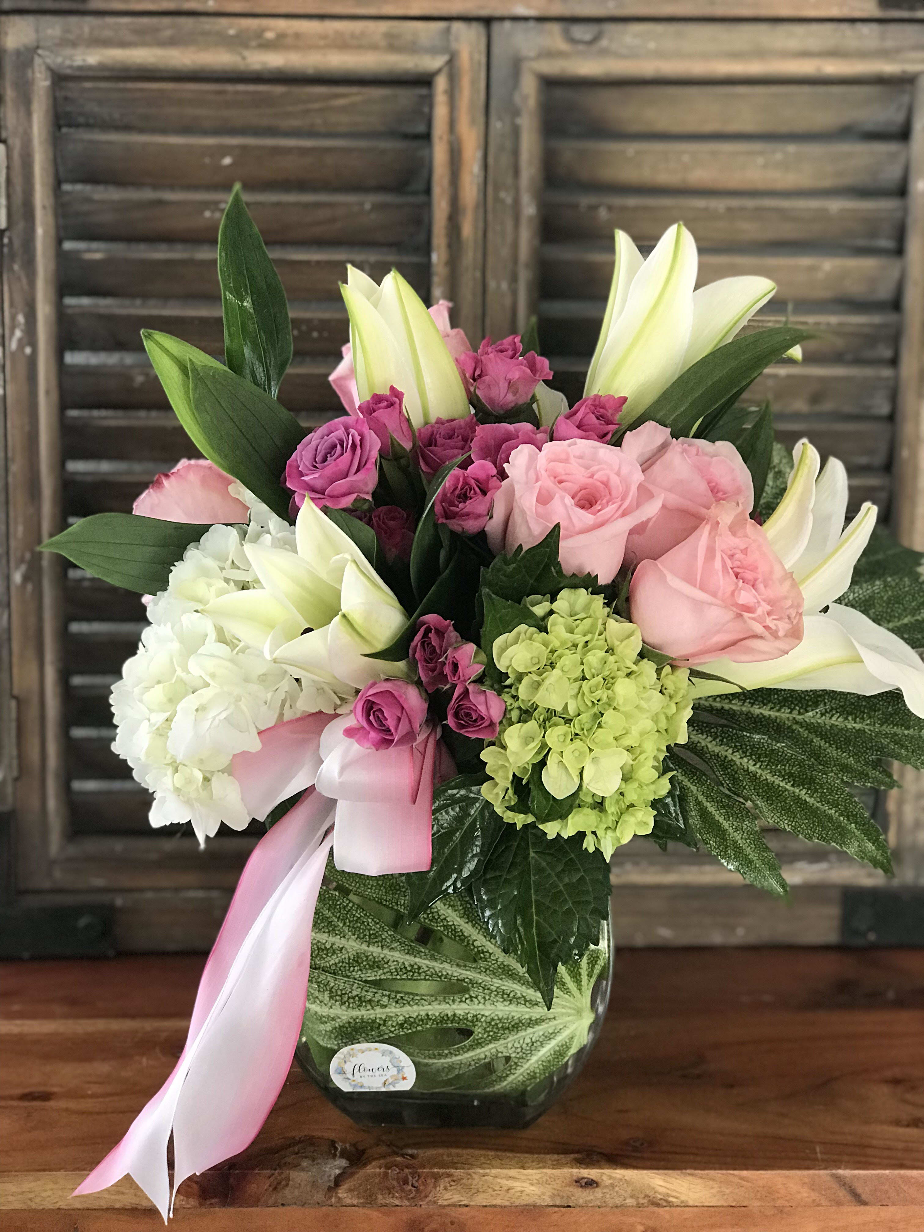 Pink and white lilies, roses, and hydrangeas in a clear glass vase with a pink ribbon bow