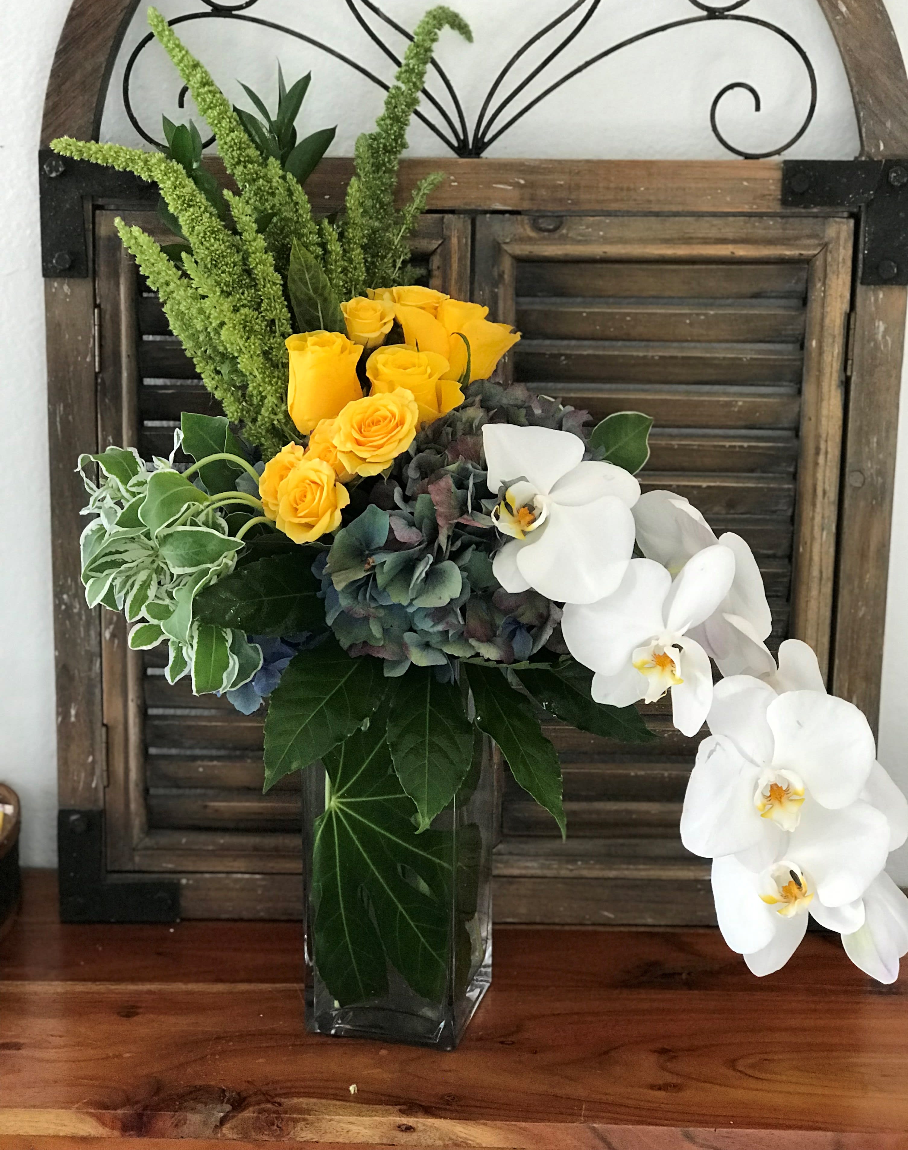 Yellow roses and white orchids in a glass vase with mixed greenery