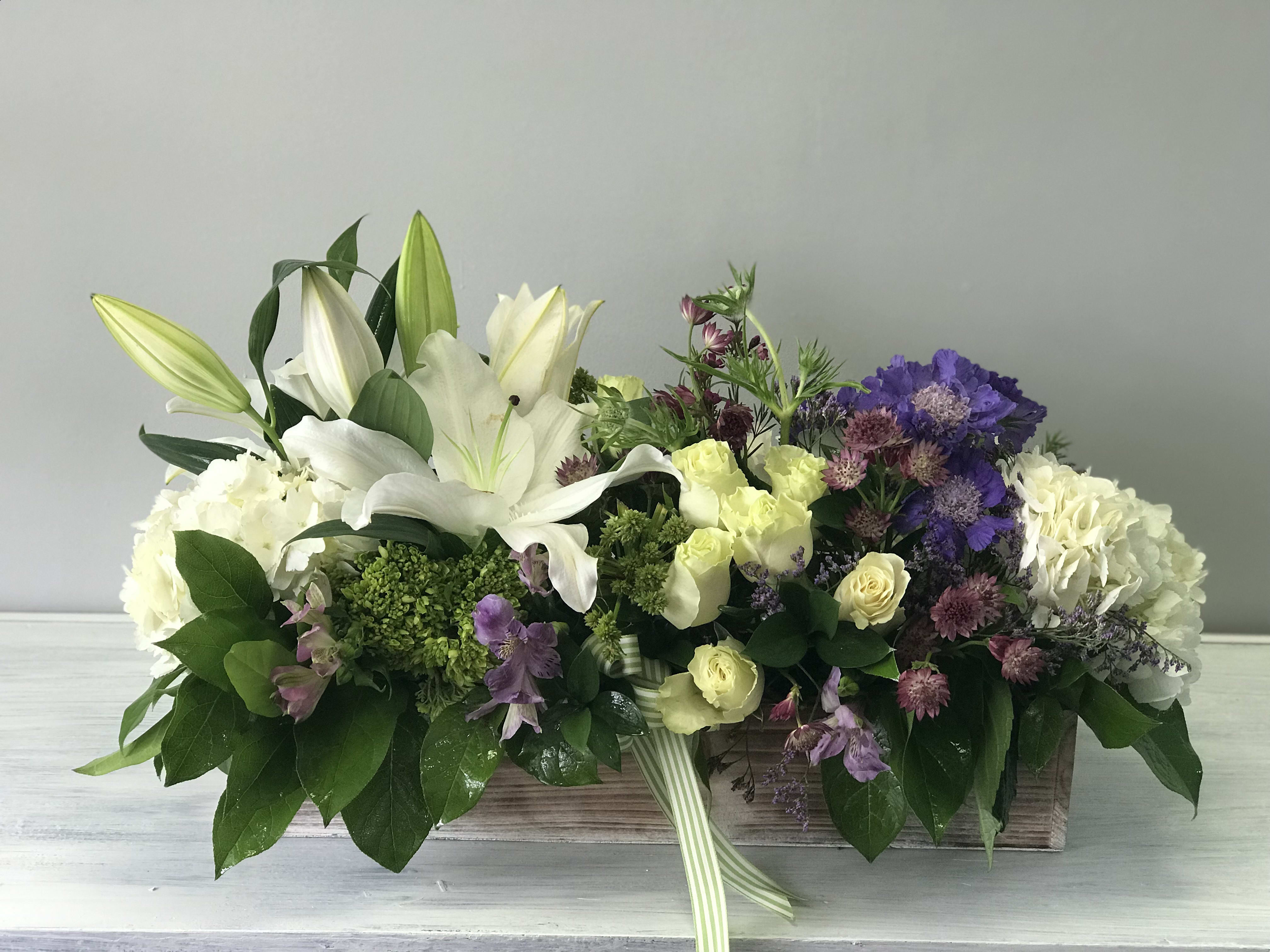 Low floral centerpiece with white lilies, cream roses, and purple blooms in a wooden box
