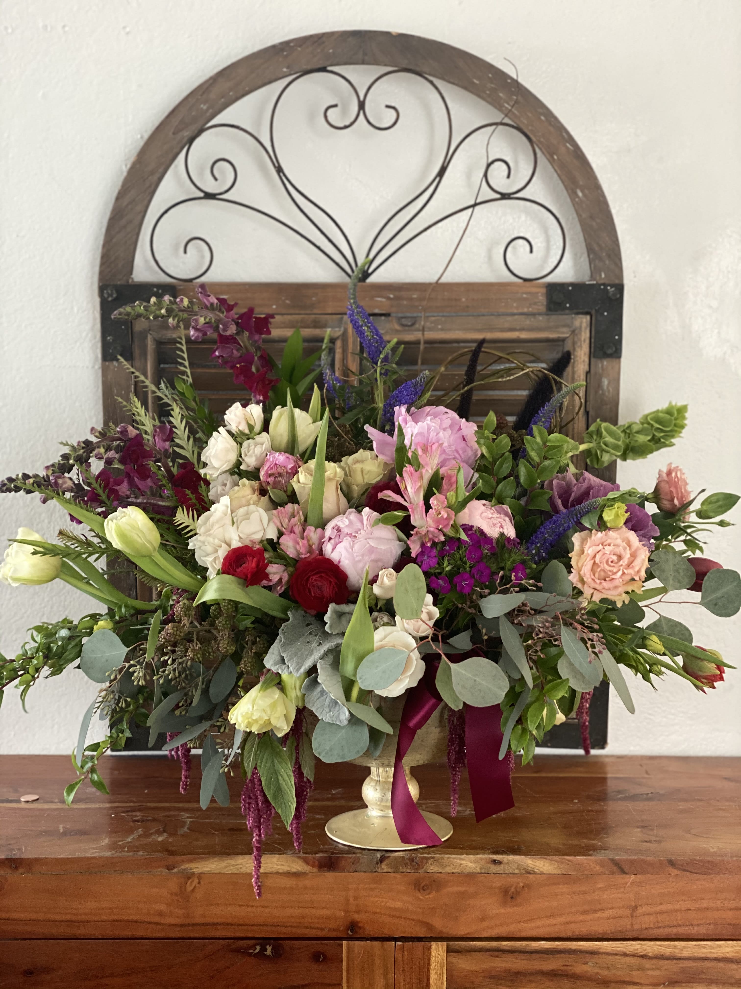 Large mixed flower arrangement in a gold pedestal vase with burgundy ribbon