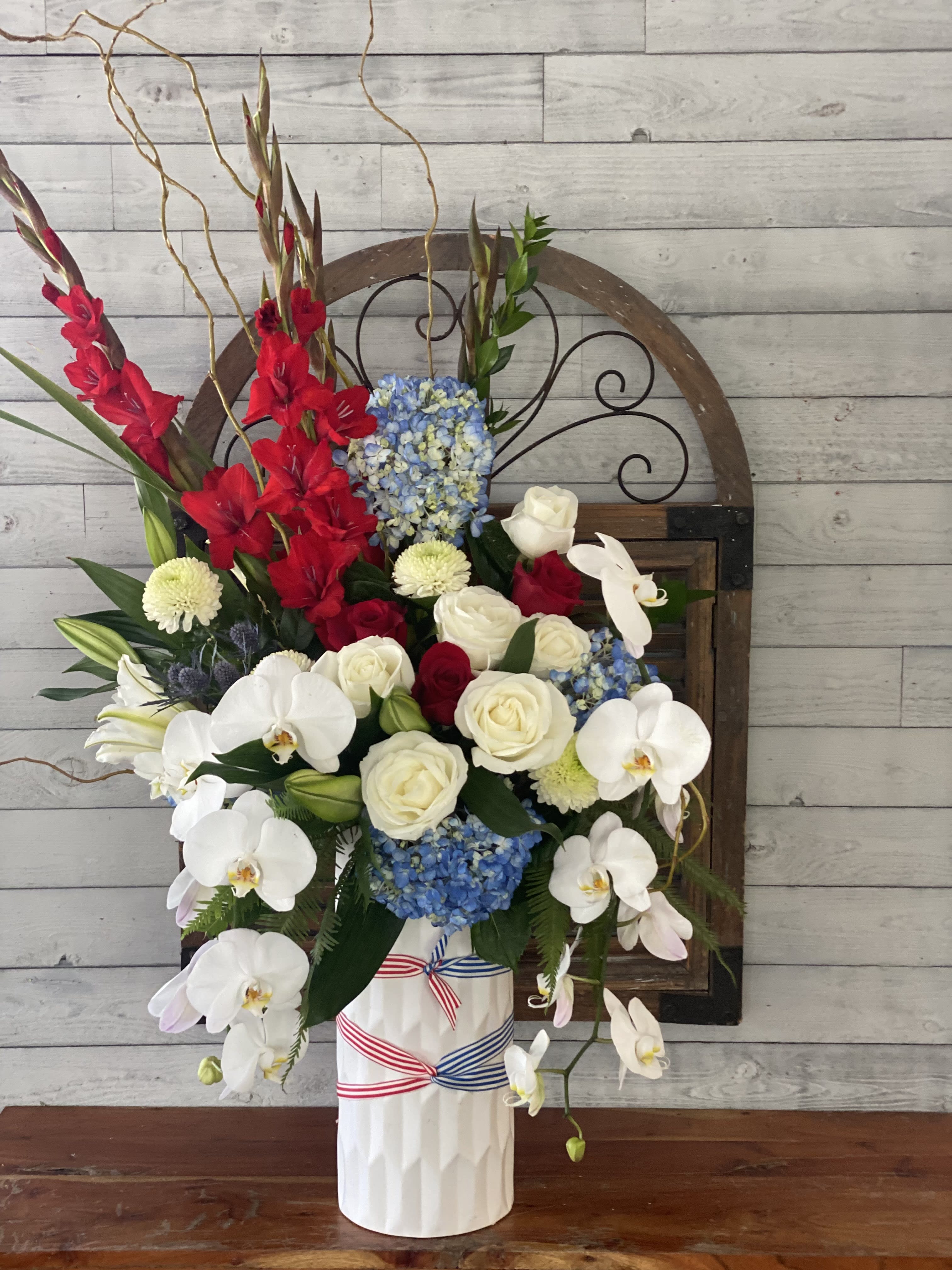 Tall floral arrangement with red gladiolus, white orchids, and blue hydrangeas in a white vase