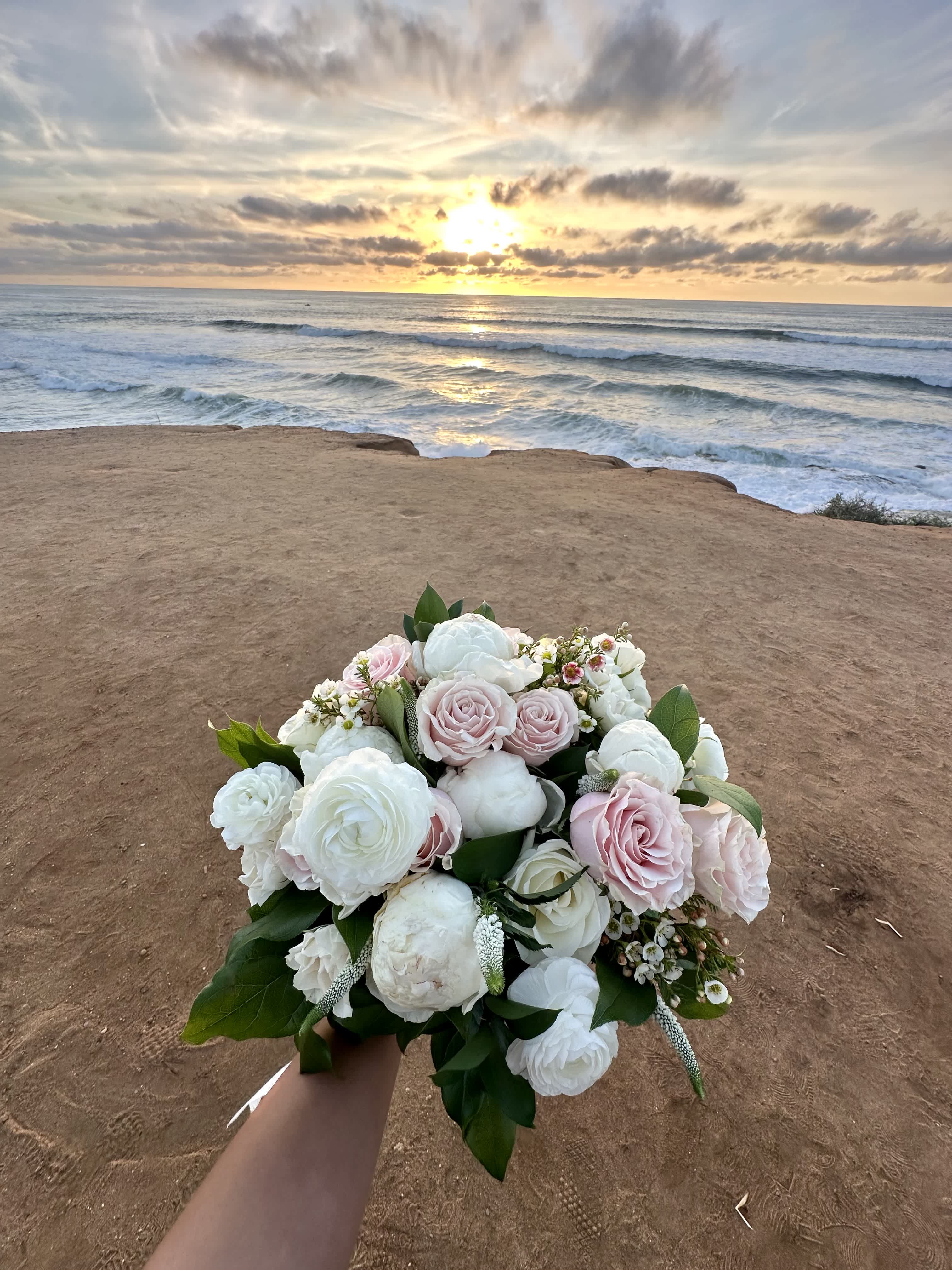 Bridal bouquet of white and blush roses and ranunculus held by the ocean at sunset