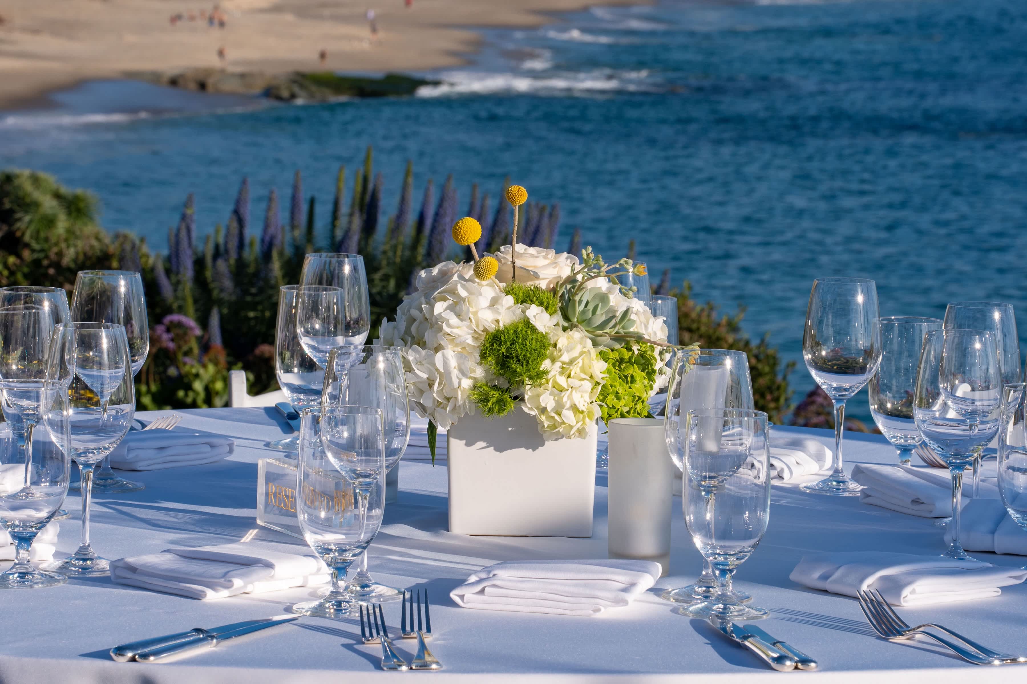 White floral centerpiece in a square vase on a set table by the water