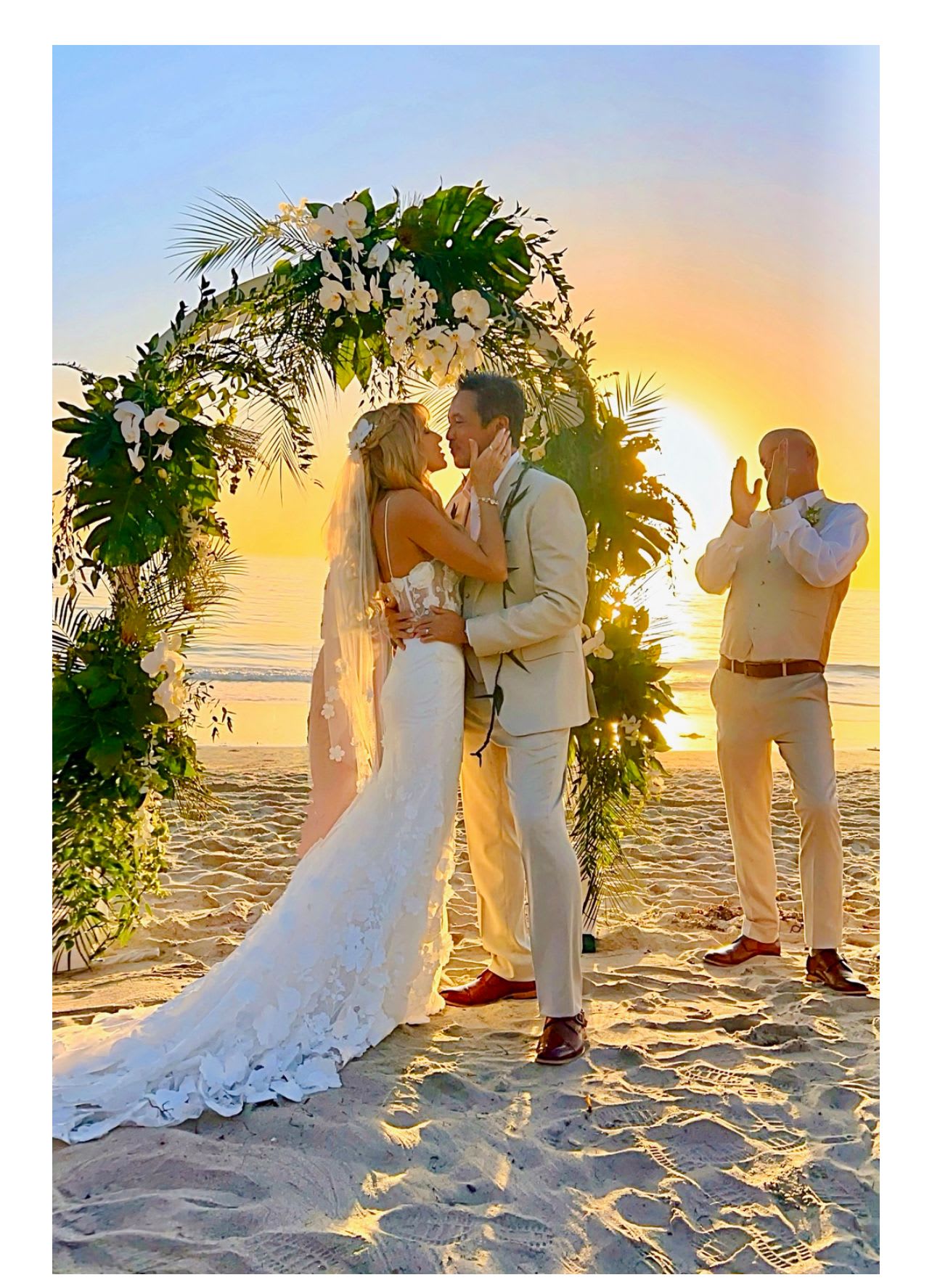 Bride and groom kissing under a floral arch on a beach at sunset