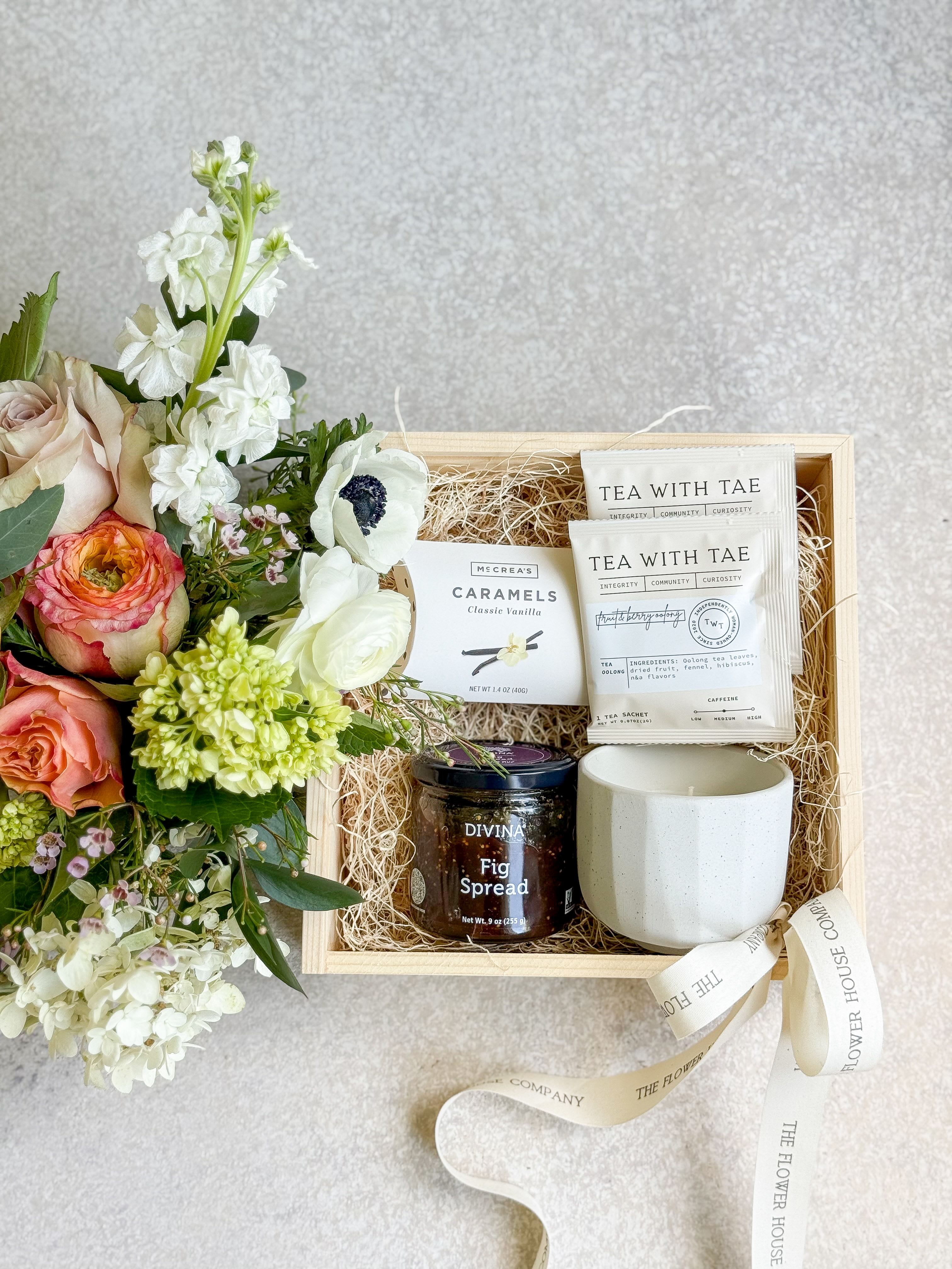 Floral gift box with roses and white blooms beside tea, caramels, fig spread, and a mug