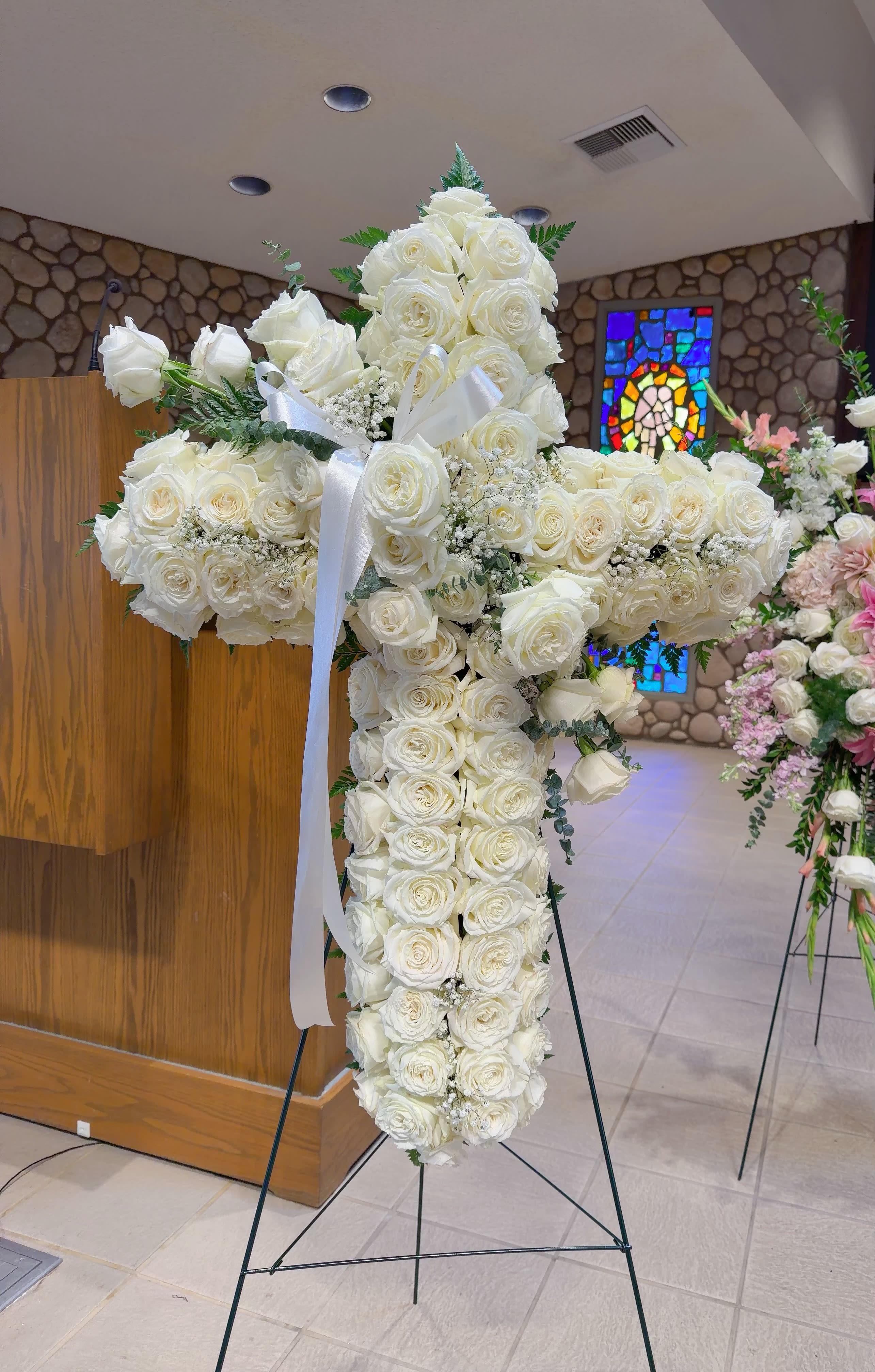 White rose cross-shaped funeral spray on a stand with a white ribbon