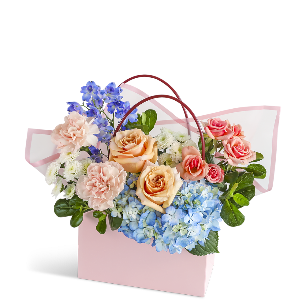 Sweet Juliette Tote - Sweet Juliette Tote features roses, hydrangea, carnations, delphinium, and premium foliage arranged in a blush pink keepsake tote. The soft mix of peach, blue, and creamy tones creates a fresh, charming design with a sweet garden feel. Bloom varieties may vary slightly with availability while the overall style remains consistent.