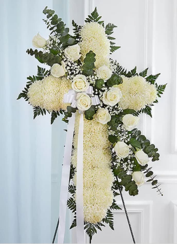 Standing Cross- White - A large standing cross crafted from densely packed white chrysanthemums creates an immediate impression of quiet reverence-perfect as a sympathy arrangement for a funeral, memorial service, or to send condolences to the family. The cross shape and white-and-green palette speak to solemnity and peace, offering the person you're honoring a dignified floral tribute that reads as both respectful and heartfelt.  Clusters of creamy white roses, small white filler blooms, and rounded greenery are artfully arranged along one side of the cross to add depth, soft texture, and visual movement against the dark green foliage outline. The arrangement sits on a sturdy standing easel and is finished with a white ribbon bow and long streamers at the center, lending a formal, traditional touch that holds up well in procession or display.  Choose this piece when you want a meaningful, faith-forward expression of sympathy that lets the bereaved feel seen and comforted. The cross arrives ready to stand on site; minimal care is required-keep it in a cool, sheltered location away from direct sun or heat until the service.