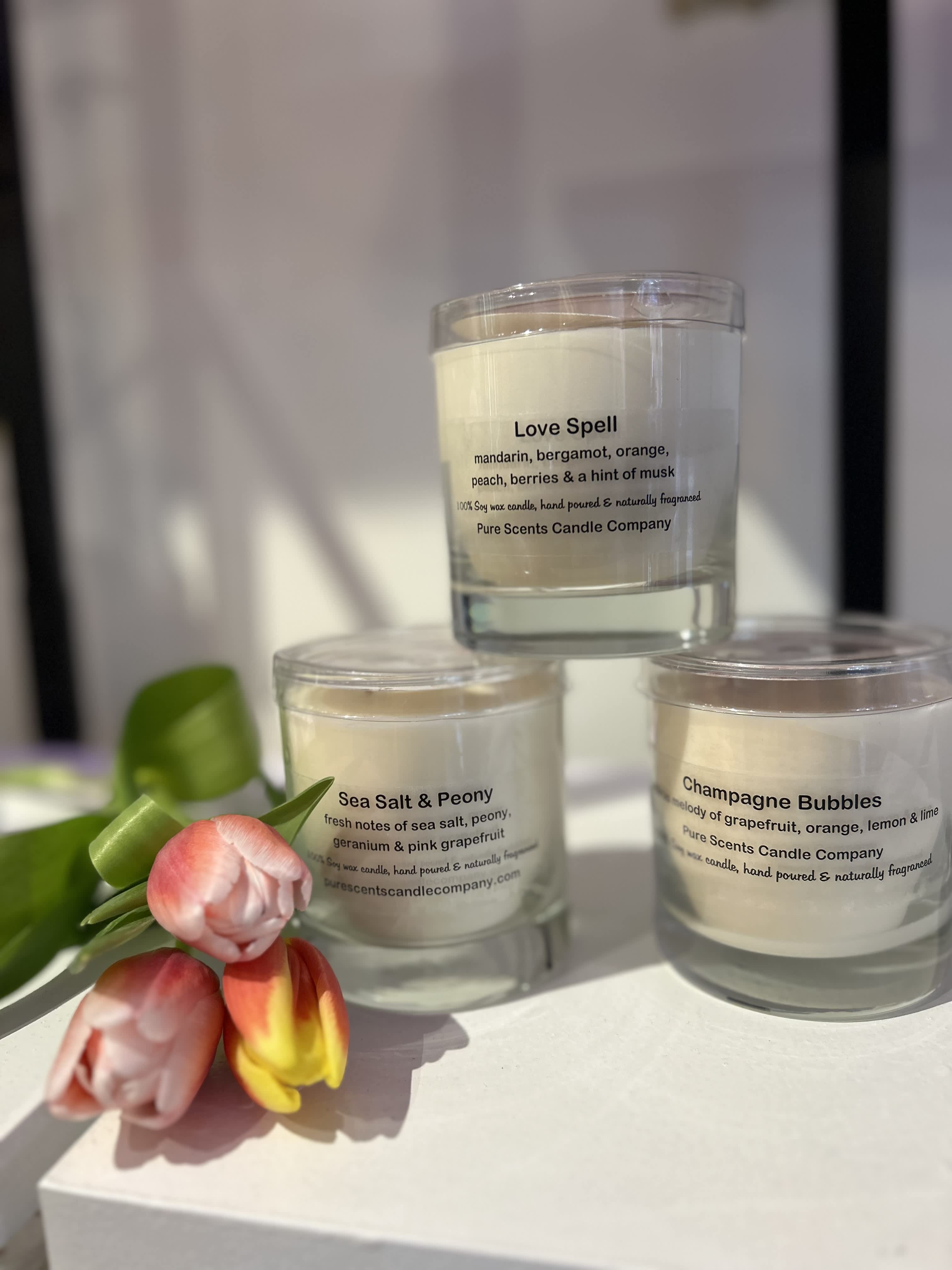 Set of three scented glass candles with printed labels, styled beside orange and yellow tulips.