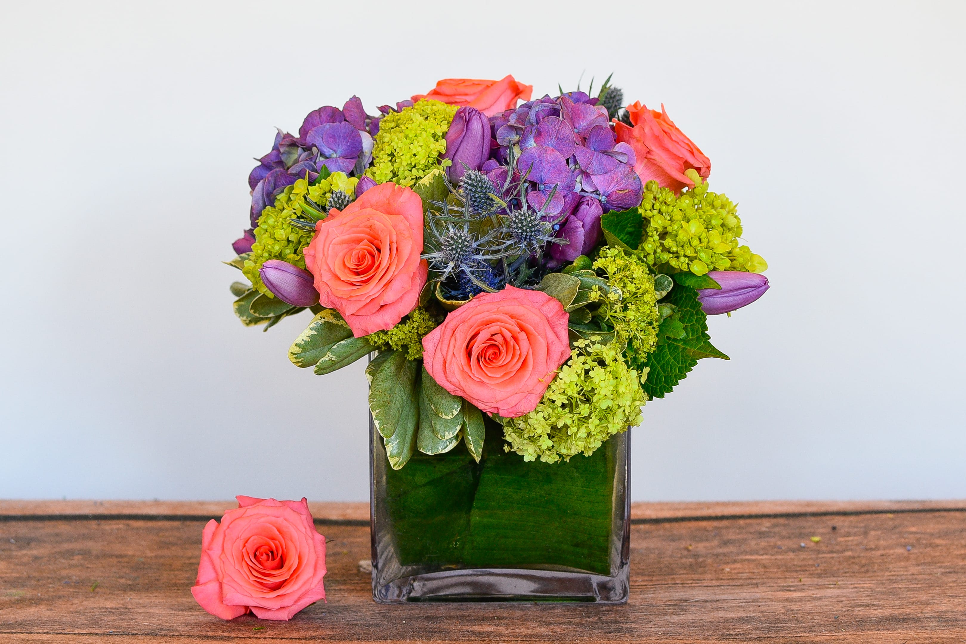 Short arrangement of coral roses, purple blooms, and green hydrangea in a square glass vase with a leaf wrap