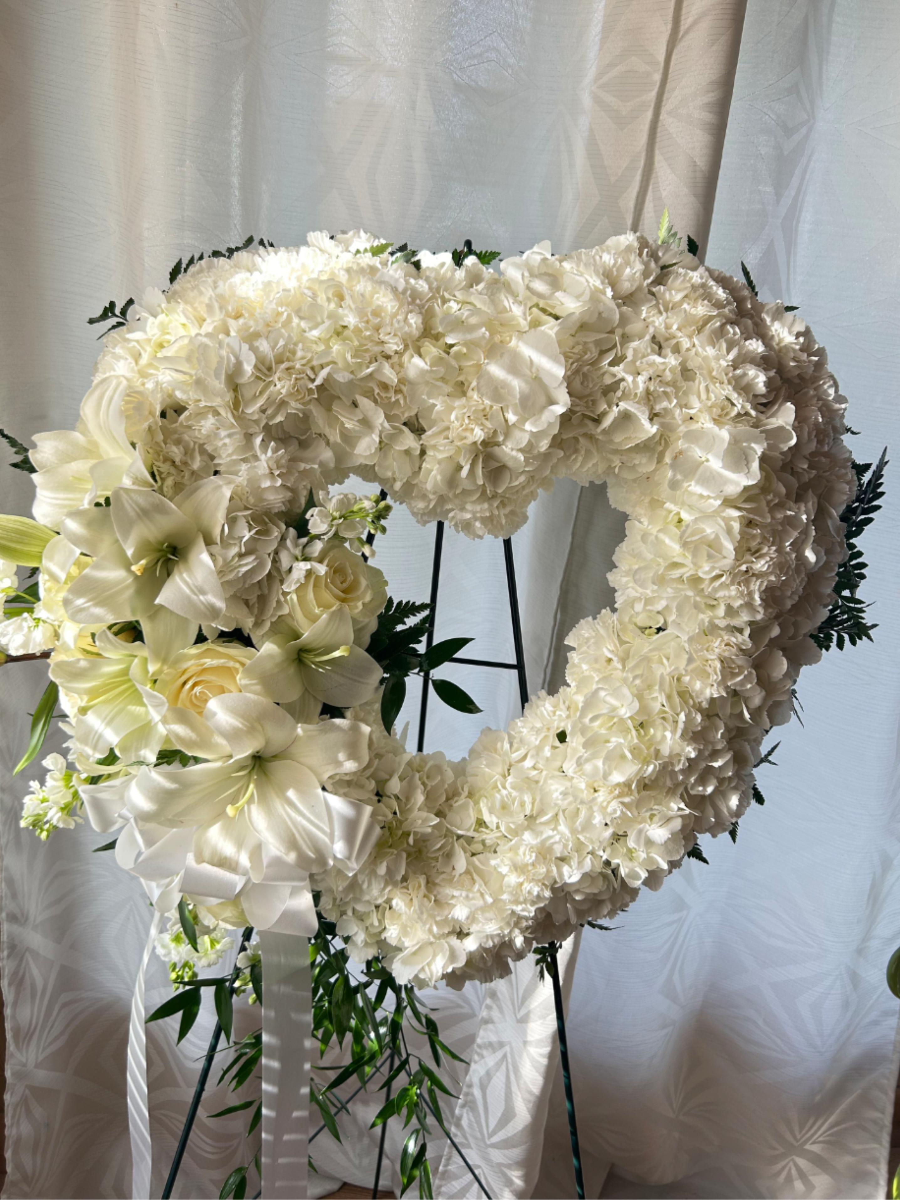 Heavenly white heart  - This elegant heart-shaped wreath is a timeless expression of love and remembrance. Designed with lush white hydrangeas, it is beautifully accented with soft white lilies and roses, delicate filler flowers, and fresh greenery. A graceful satin ribbon bow adds a finishing touch, enhancing its classic and peaceful appearance. This arrangement symbolizes purity, devotion, and eternal love, making it a meaningful tribute for memorial services, celebrations of life, or heartfelt expressions of sympathy. 
