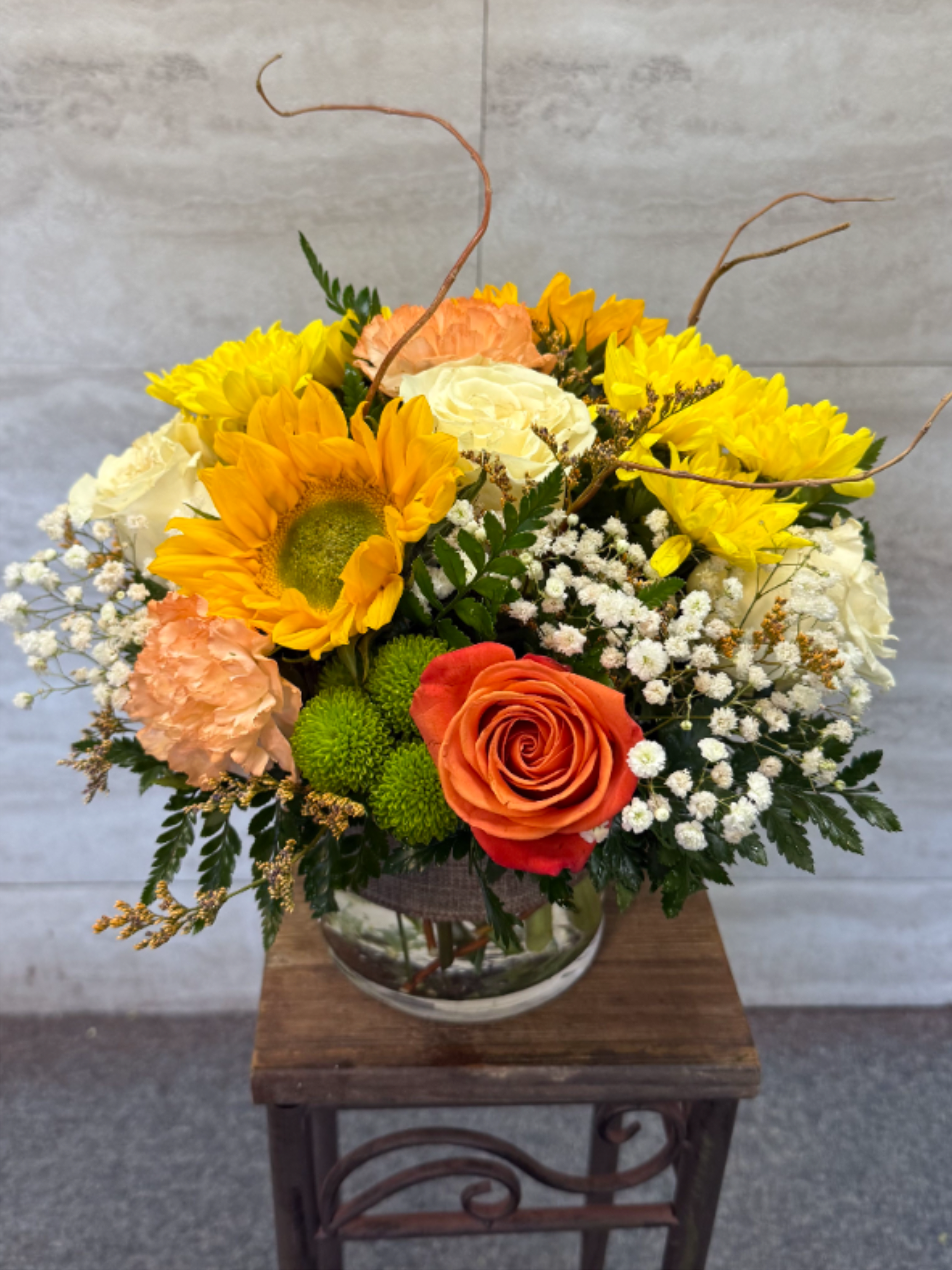 Golden Hour Glow - (ALL-ROUNDED) A cheerful mix of sunflowers, creamy roses, peach carnations, and a vibrant orange rose, accented with baby’s breath and lush greenery. Designed in a clear glass vase, this bright and happy arrangement brings warm sunset vibes into any space. 
