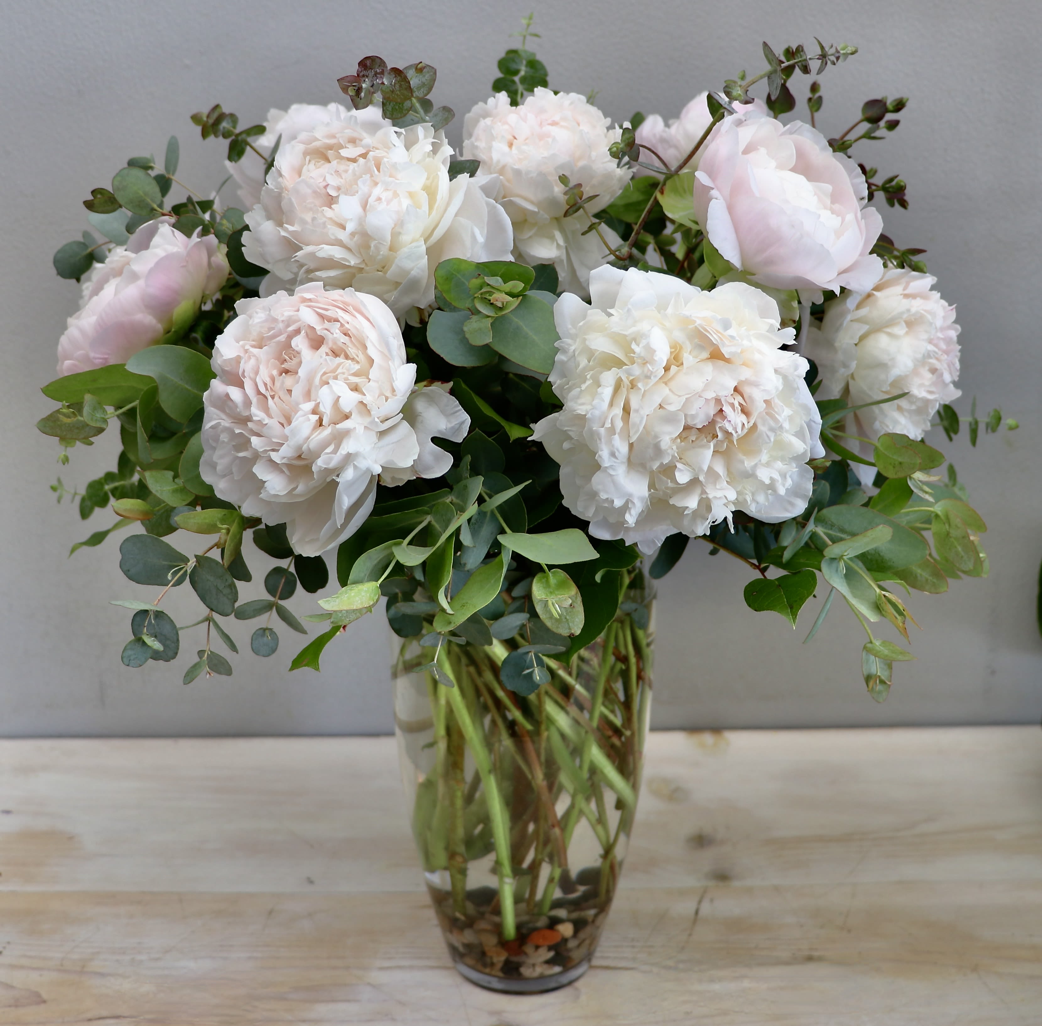 Tall Blush Peony - Glendale Flowers - Layers of delicate light pink peonies spill over the rim of a textured, tall glass vase for a look that is both timeless and tactile. This arrangement balances the airy softness of spring blooms with the clean, rhythmic lines of contemporary glasswork.