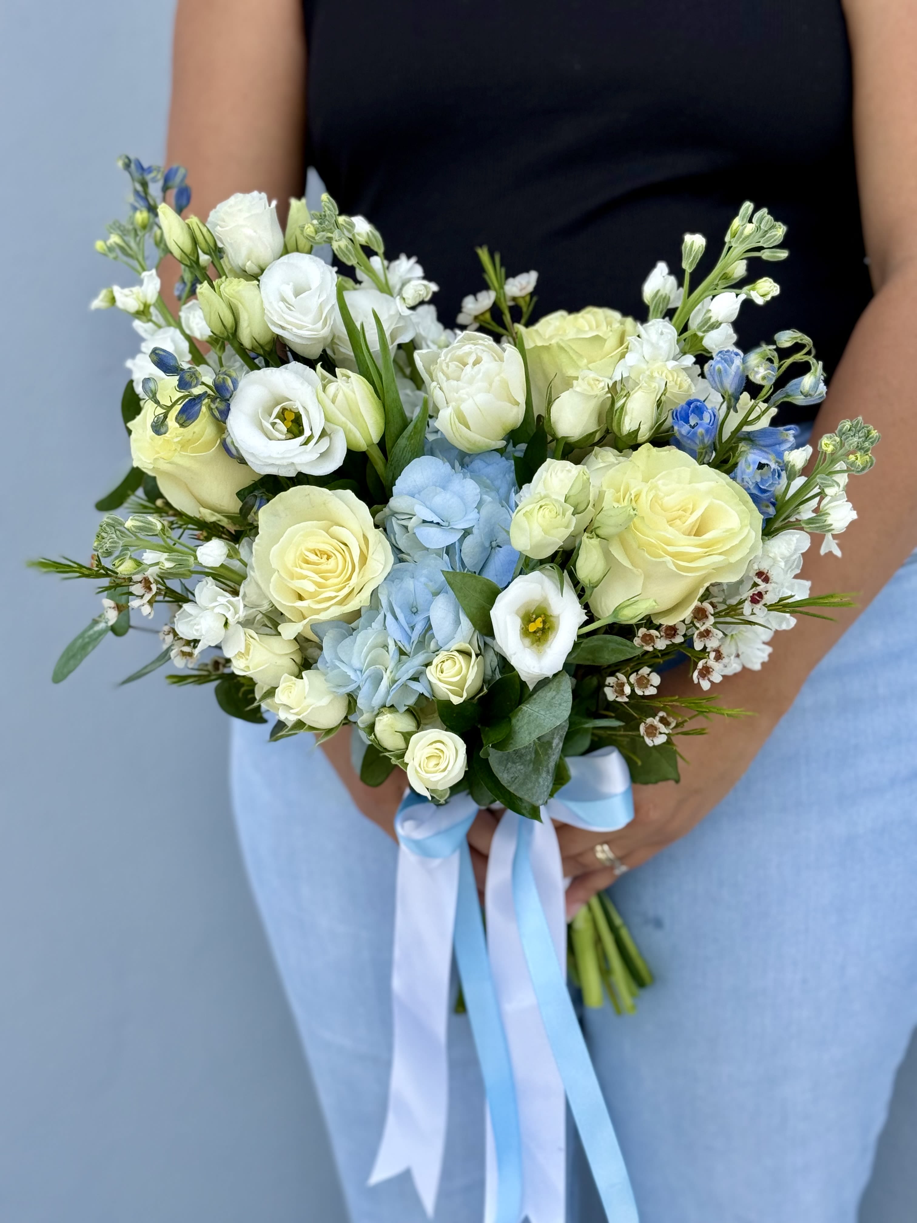 Handheld bouquet of white and pale yellow flowers with blue accents and ribbon