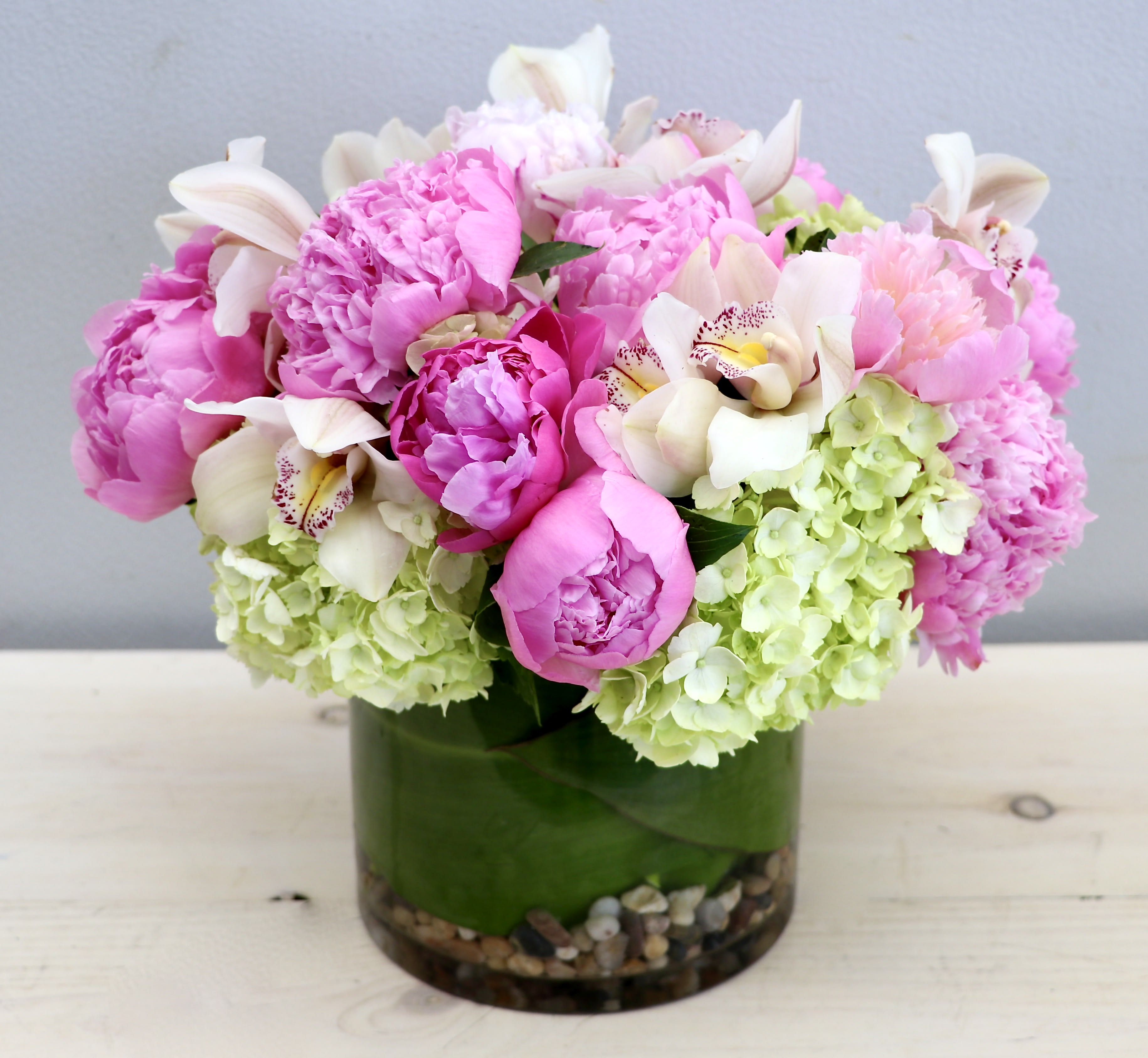 Compact arrangement of pink peonies, white orchids, and green hydrangeas in a glass cylinder vase